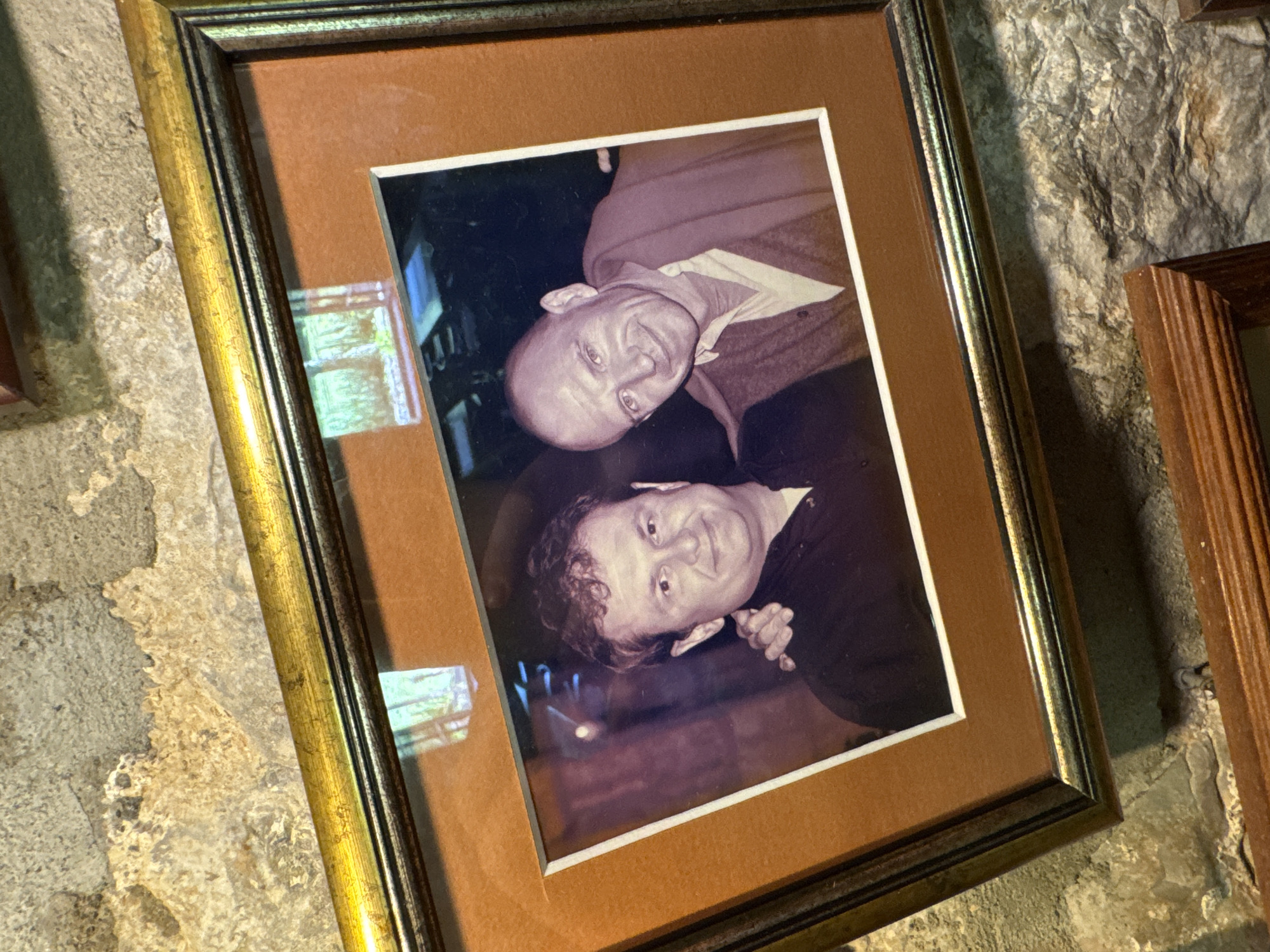 A framed photograph, tilted slightly, shows two men smiling at the camera. The man on the left has curly brown hair and is wearing a dark shirt. The man on the right is bald and wears a light-colored shirt with a cardigan draped over his shoulders. They appear to be indoors, with a blurry background suggesting a dimly lit environment. The photograph is framed in a gold and dark-colored frame with a brown mat. The frame is leaning against a rough, textured stone wall. This scene is located in the city of Morinj, Montenegro.