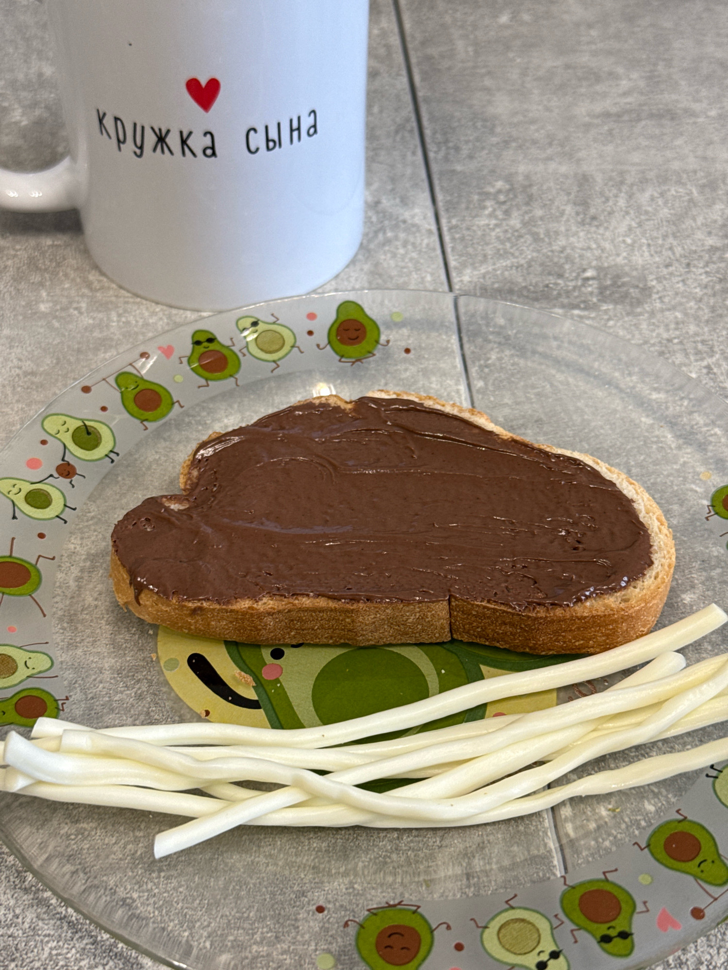 A slice of bread, spread with chocolate-hazelnut spread, rests on a clear glass plate with avocado illustrations. Next to the bread, on the plate, are several pieces of string cheese. In the background, a white mug with a red heart and Russian text that reads "кружка сына" (son's mug) is visible. The scene appears to be on a tiled countertop in Lyubertsy, Russia.