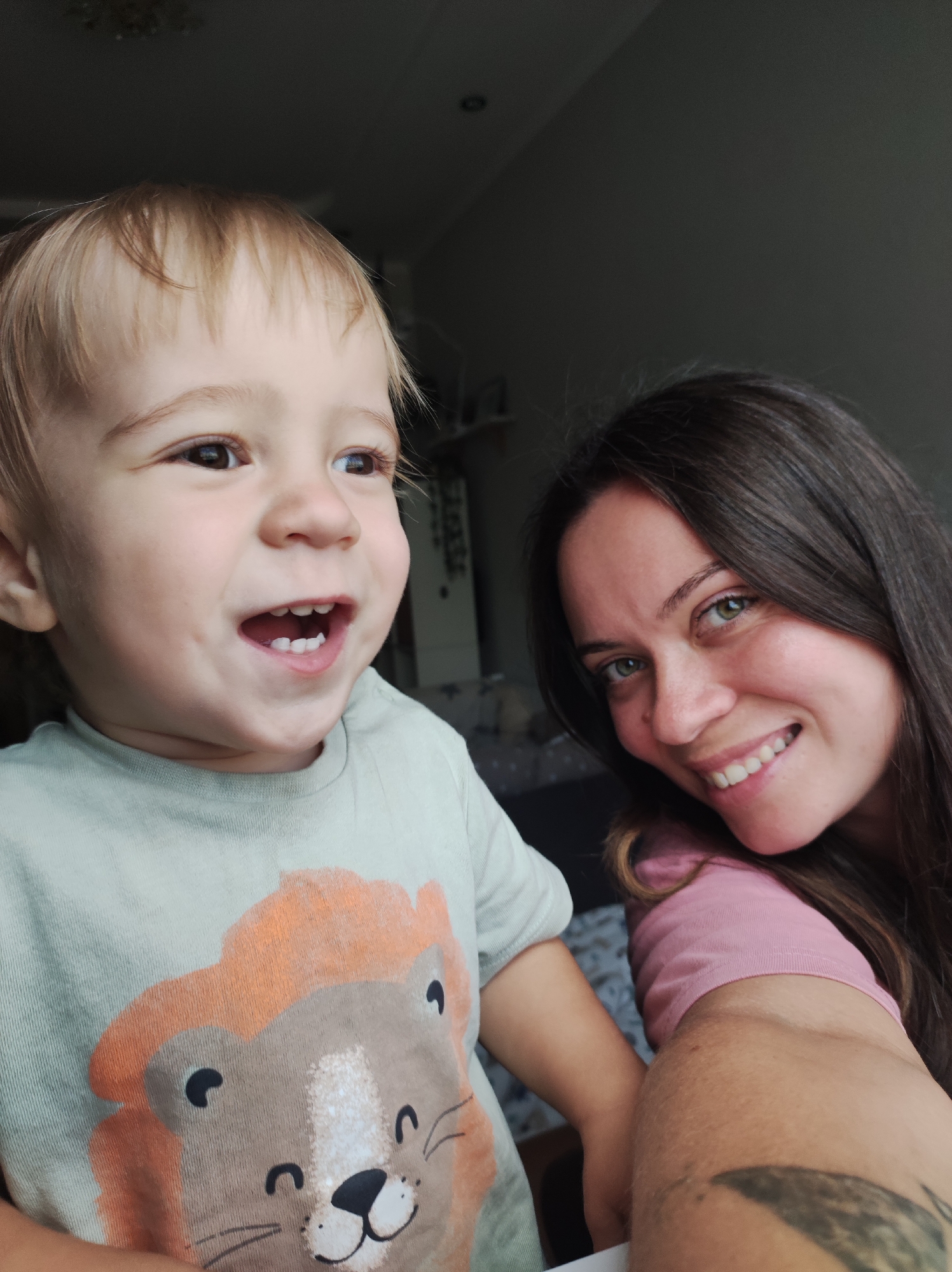 A selfie shows a young child with blonde hair and a light green t-shirt featuring a lion graphic, looking to the left and smiling widely with mouth open. Next to the child, a woman with long dark hair and green eyes smiles at the camera, wearing a pink t-shirt. The background is dark and out of focus, suggesting an indoor setting, possibly a home, in Chaikovsky, Russia.