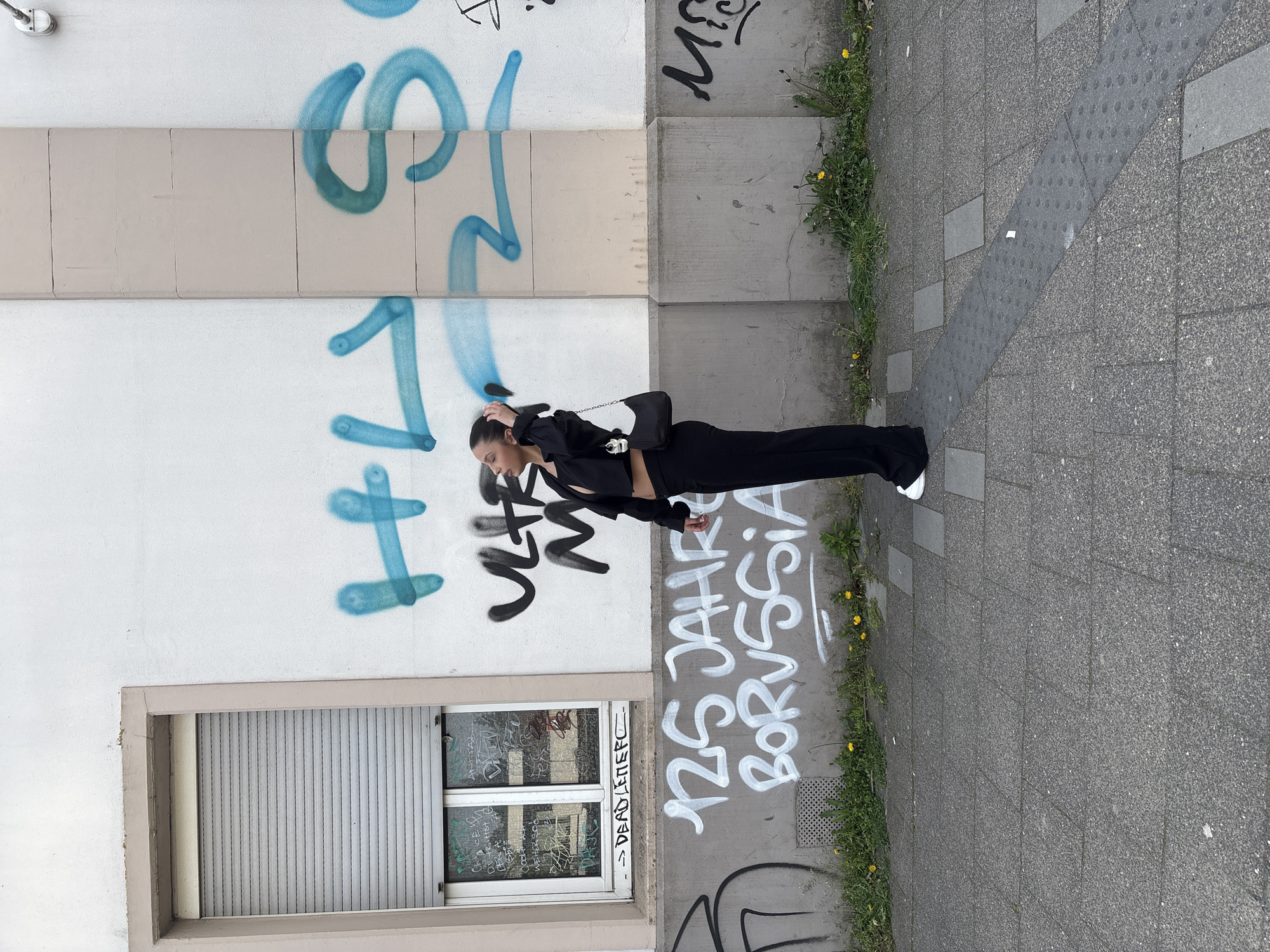 A woman is standing on a sidewalk next to a wall covered in graffiti. The woman is wearing a black outfit and white shoes, and she has a black purse over her shoulder. The wall is white with blue and black graffiti, including the words "125 Jahre Borussia." There is also a window with graffiti on it, and a small patch of grass with dandelions growing next to the sidewalk. The location is Mönchengladbach, Germany, as indicated by the graffiti "125 Jahre Borussia," which references the German football club Borussia Mönchengladbach.
