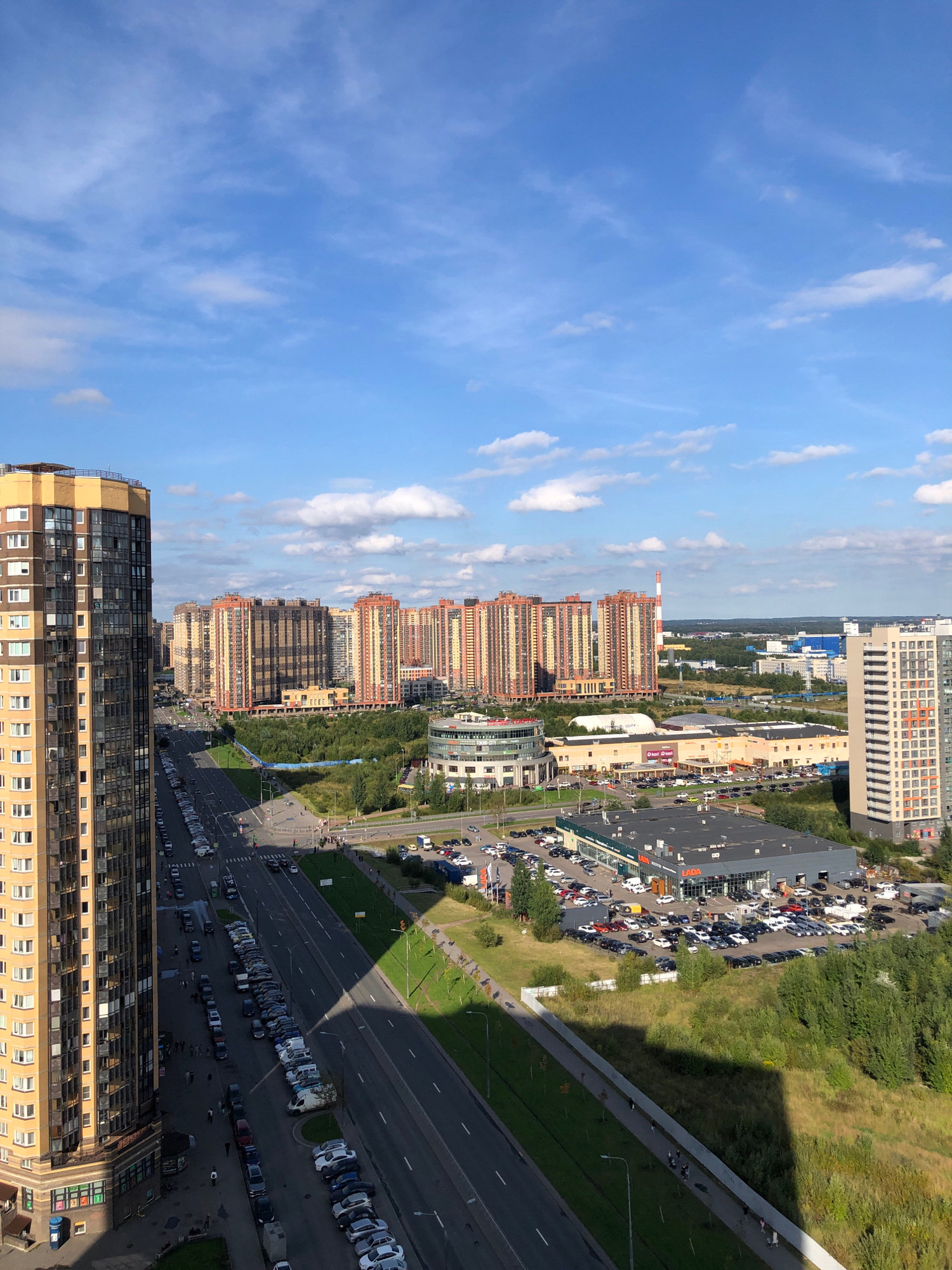 This is an aerial, high-angle, daytime, full shot of a suburban cityscape in Lavriki, Russia, under a blue sky with scattered white clouds. The foreground features a tall, multi-story residential building with a yellow and dark-paneled facade. To the right of this building is a multi-lane asphalt road lined with parked cars on both sides. Beyond the road, a long, strip mall-like building with a dark gray roof, labeled "LADA," is visible, surrounded by a large parking lot filled with vehicles. Further in the distance, several large, mid-rise residential complexes with orange and beige facades dominate the landscape. Greenery, including trees and grass, is interspersed throughout the scene, particularly in the areas between buildings and along the roadside. Several figures, appearing as small silhouettes, are visible walking along a pathway adjacent to the road. The overall scene depicts a modern urban development with residential and commercial areas.