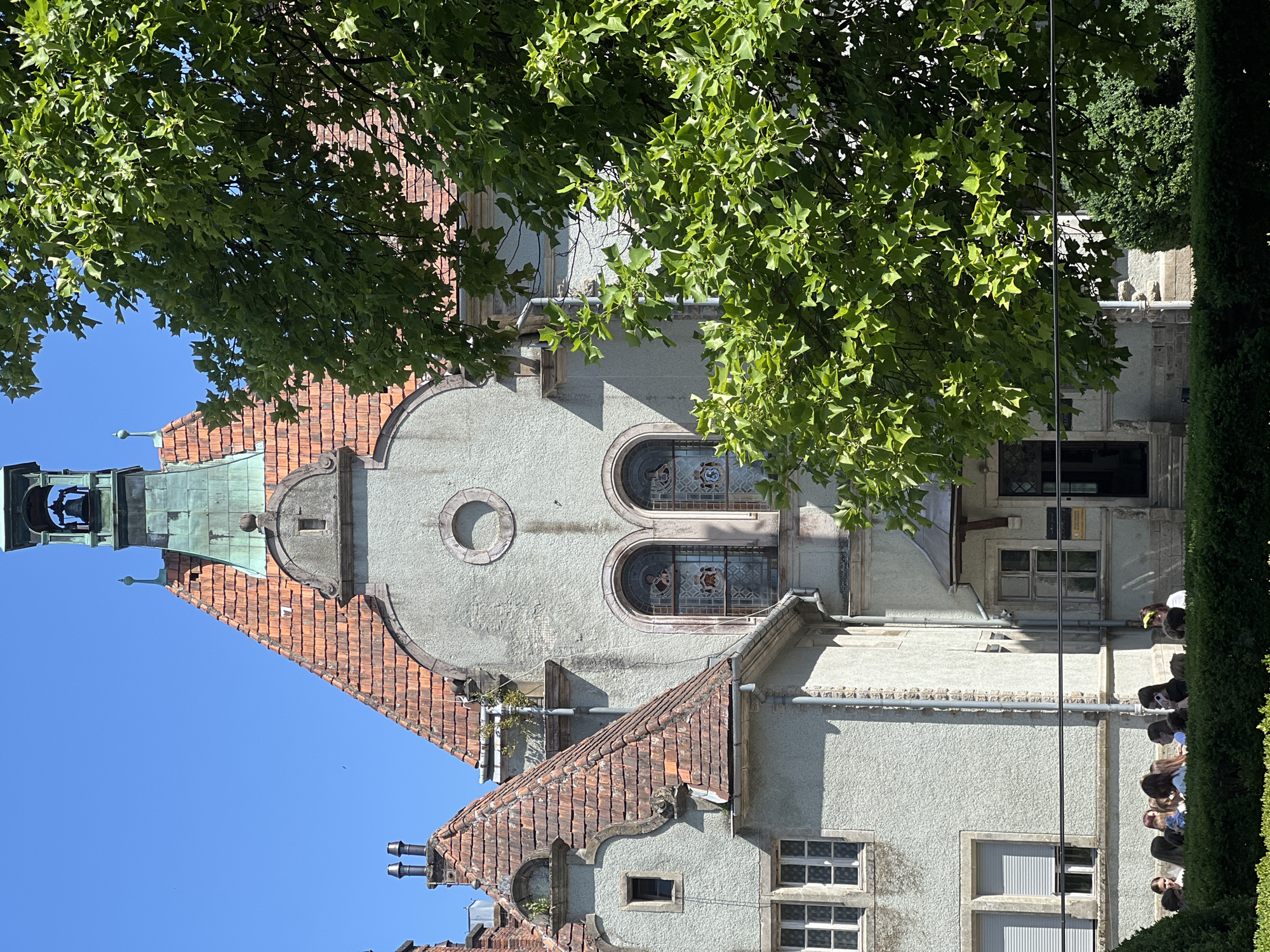 The image shows a detail of a grand building with red tiled roofs and a bell tower. The building's facade is light gray and features arched stained-glass windows and a circular window. Lush green trees with large leaves partially obscure the view of the building. In the foreground, a group of young people are gathered behind a hedge, appearing to be on a school trip or excursion. The sky is a clear, bright blue. The scene is indicative of a sunny day in an outdoor setting, possibly a park or a historic site. The architectural style suggests an older, possibly European, building. The mention of Karpaty, Ukraine, places the scene in a mountainous region known for its natural beauty and historical sites.