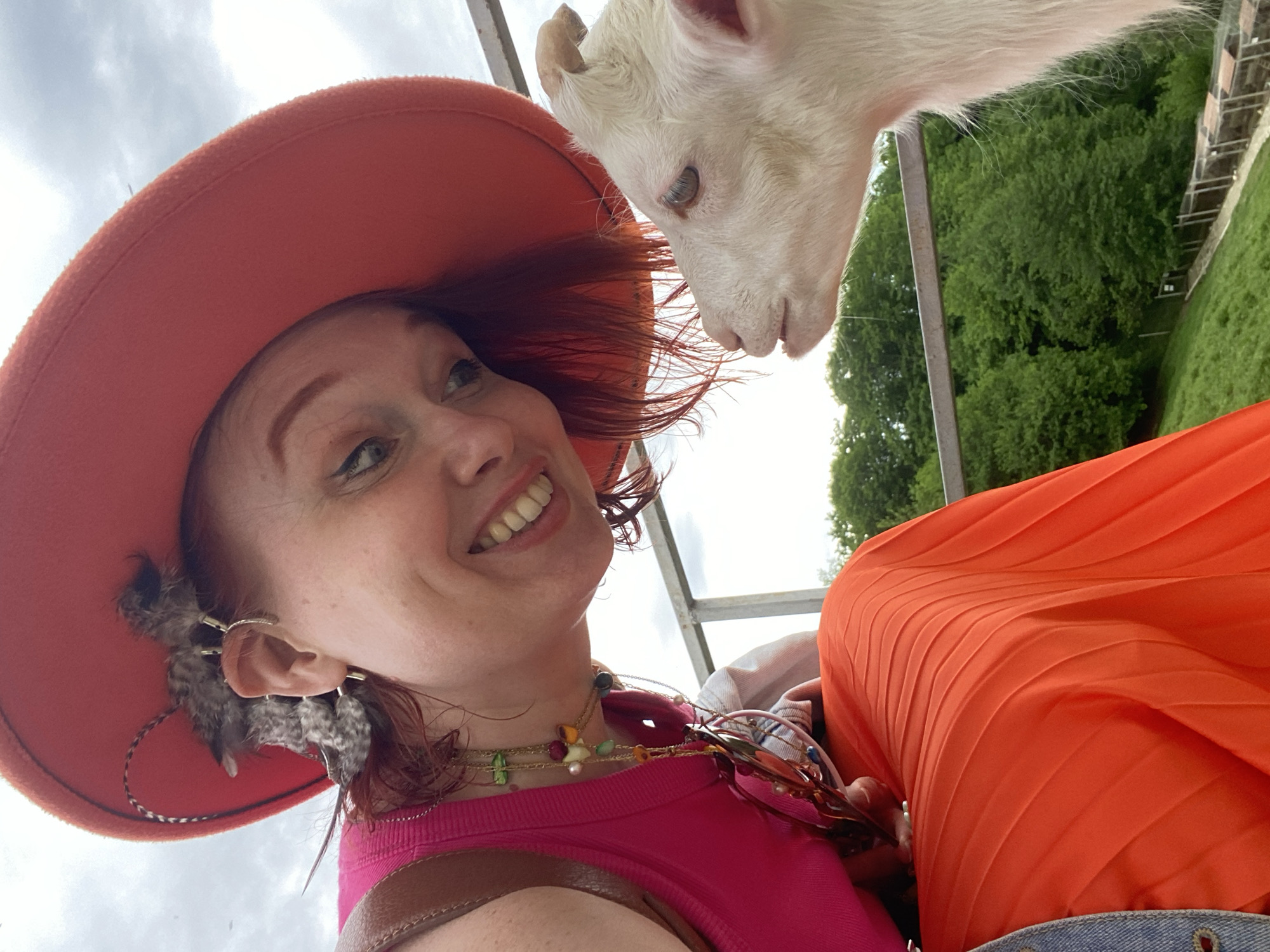 A person wearing an orange hat, a pink top, a necklace, and earrings with feathers is smiling while looking at a white baby goat. The goat is leaning over a metal railing, with green trees and a grassy hillside visible in the background.