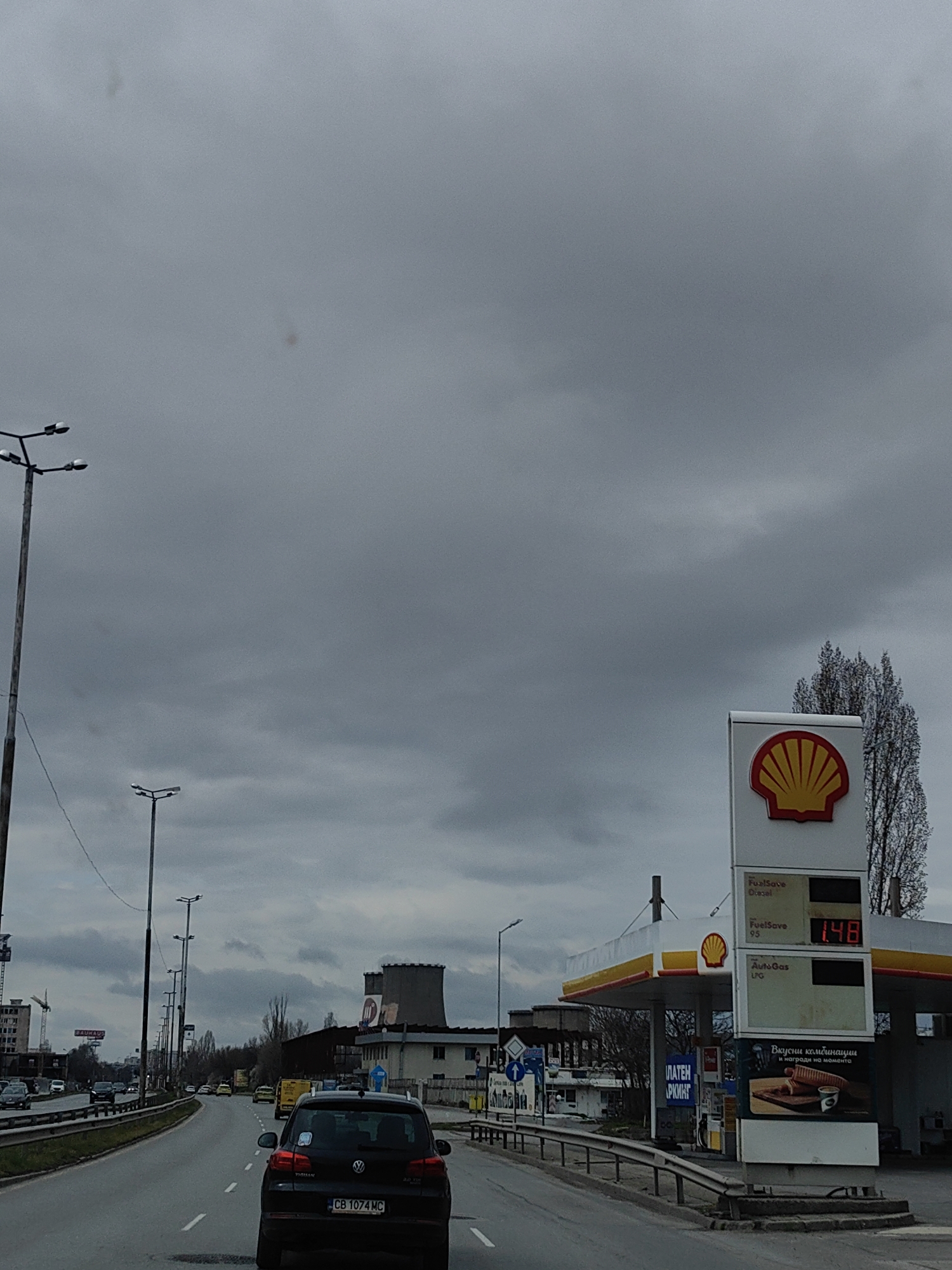 The image shows a road scene in Sofia, Bulgaria on an overcast day. In the foreground, a black Volkswagen Tiguan with the license plate "CB 1074 MC" is driving on the road. To the right of the road is a Shell gas station, with the Shell logo prominently displayed. The gas price sign shows a price of "1.48" for FuelSave 95. Streetlights line the road on the left. Further down the road, in the middle ground, is a large, cylindrical industrial structure. The sky is filled with dark, gray clouds. Other cars can be seen on the road in the distance. A BAUHAUS sign can be seen in the distance on the left side.