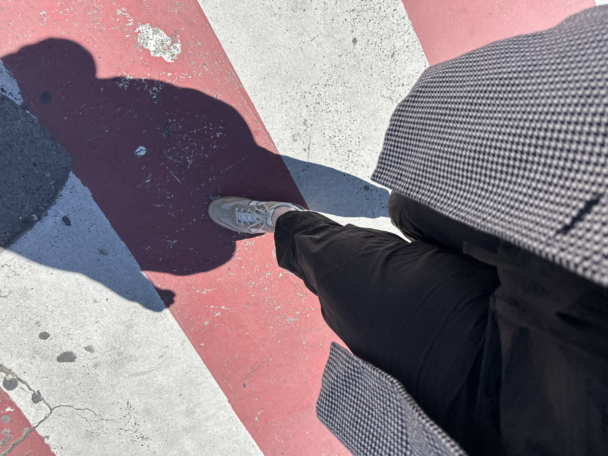 An overhead shot depicts a person standing on a crosswalk marked with red and white stripes. The person is wearing grey sneakers, black trousers, and a grey and black houndstooth coat. Their shadow falls across the red and white stripes. The photograph appears to be taken in Tbilisi, Georgia.