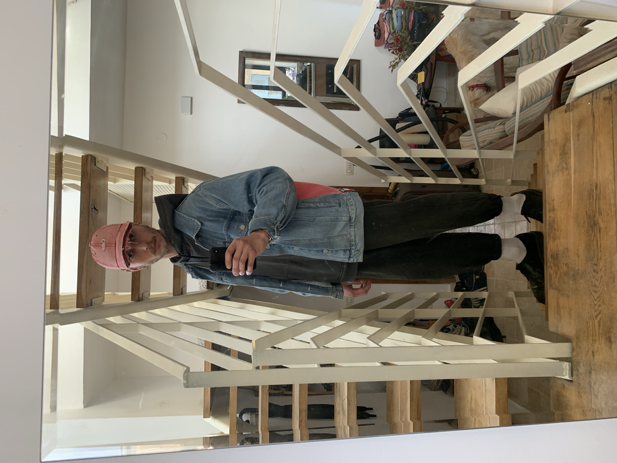 A full-body mirror selfie captures a male subject positioned on a wooden staircase within a residential interior in Tel-Aviv, Israel. The subject, appearing to be an adult male with light facial hair, is dressed in a distressed pink baseball cap displaying "Coke" in white script, a grey hooded sweatshirt, a light blue denim jacket, dark cuffed pants, white socks, and black shoes. He holds a black smartphone in his right hand, directed towards the mirror, in the act of photography. A red object, potentially a backpack strap, is partially visible behind his right shoulder.
The immediate environment features a wooden staircase with light-toned steps and a white painted metal railing characterized by a geometric pattern of diagonal and horizontal bars. The walls are white. To the subject's left in the reflection, more wooden stairs ascend, and dark objects, possibly shoes, are visible on shelves along a wall. To the subject's right, a dark wooden-framed mirror is mounted on the wall. Below this mirror, a surface holds a stack of folded textiles in various colors and a small floral arrangement with red flowers. Further to the right and lower, partially obscured by the railing, are sections of a striped upholstered armchair and a light-colored dog wearing a red collar. Miscellaneous bags, a suitcase, and other household items are present in the lower right background. Electrical outlets and a small white rectangular device are visible on the white walls.