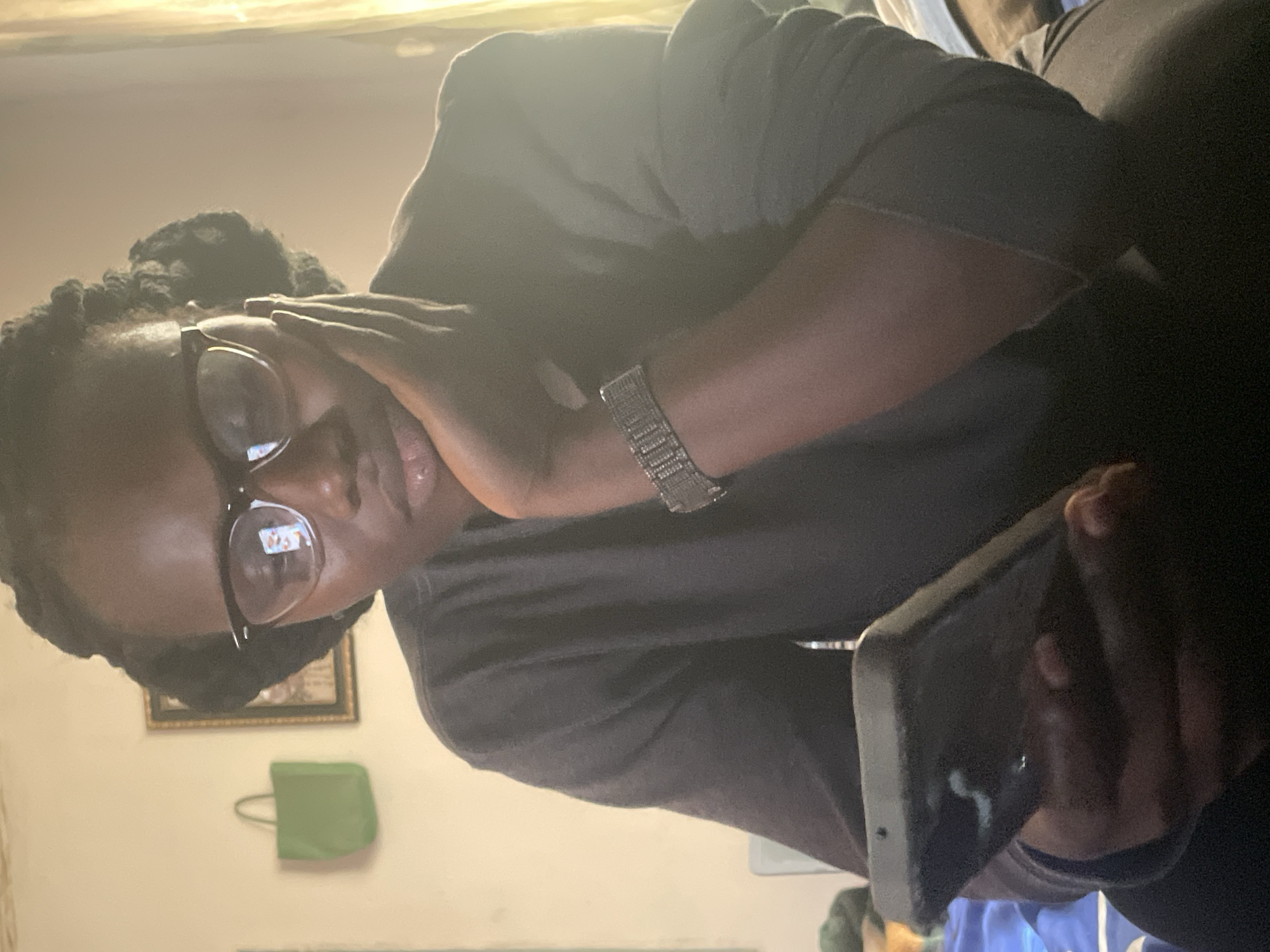 A person is holding a smartphone in their right hand, and their left hand is touching their face. The person is wearing glasses and a bracelet. A painting and a green bag are on the wall behind the person. The location appears to be indoors, but there is no indication in the picture that it is in Jos, Nigeria.