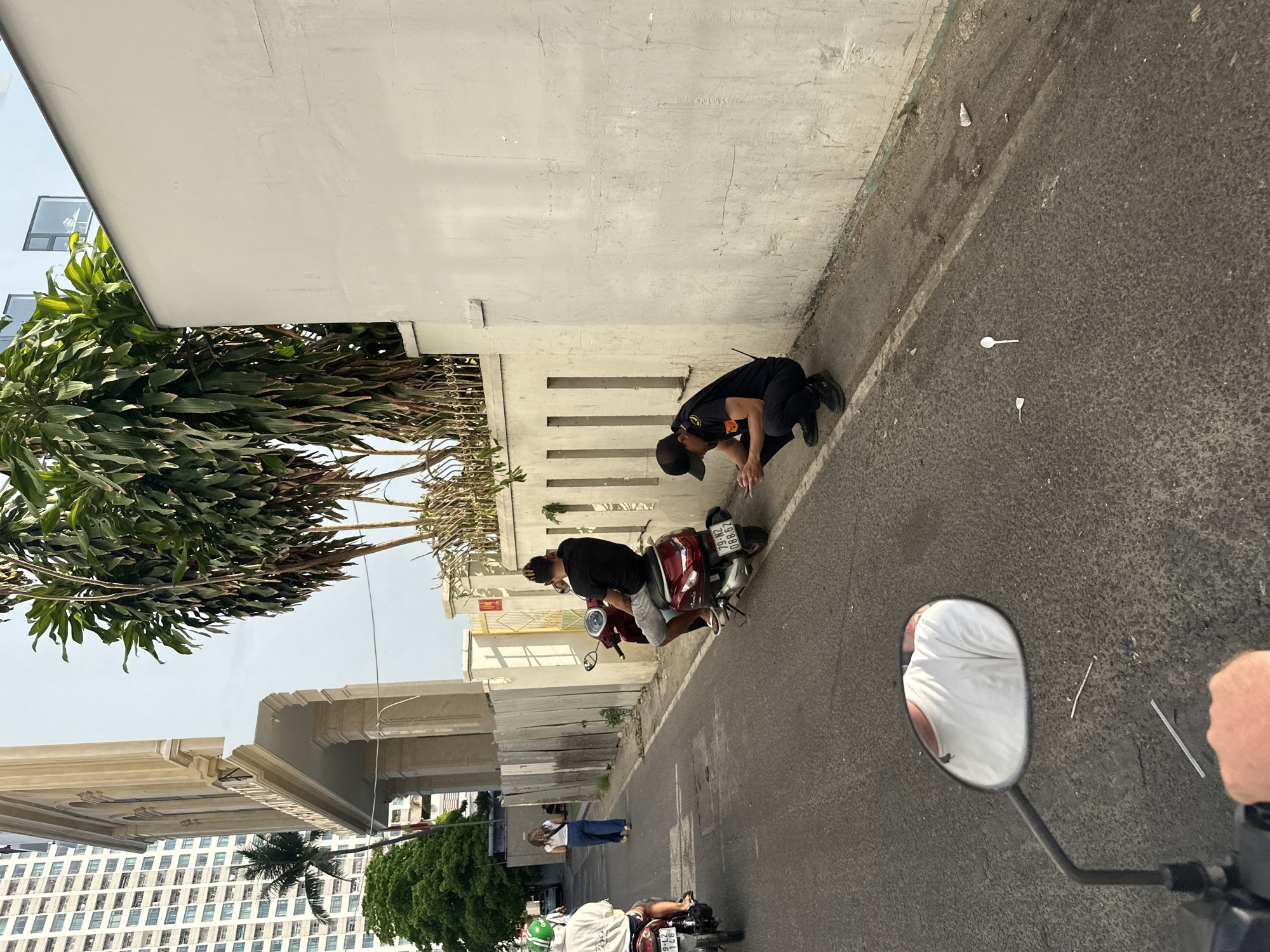 The image depicts a street scene in Bac Nha Trang Ward, Vietnam. A person in a dark uniform and baseball cap is kneeling next to a parked red scooter on the side of a street with a white wall, possibly inspecting something on the ground. A second person is on the scooter. Plastic waste is on the street. In the distance, other people are walking on the street. Buildings and trees line the other side of the street. The view is from the perspective of someone on another scooter, with a side mirror visible in the foreground.