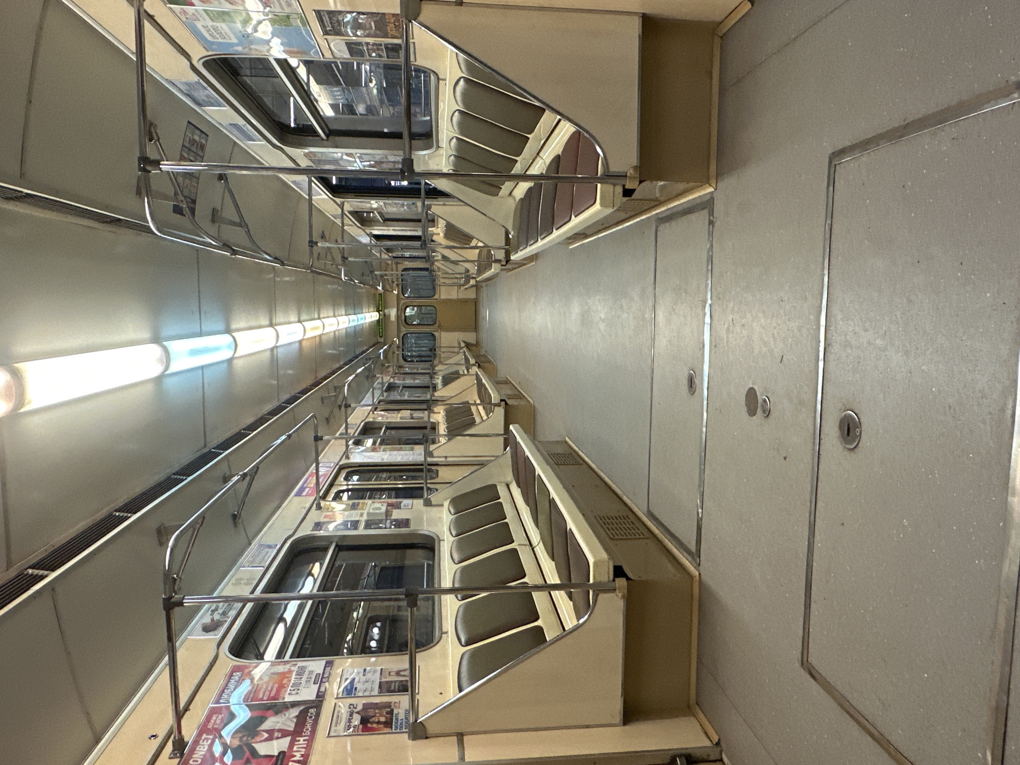 The image shows the interior of an empty subway train car, likely on the Minsk Metro in Belarus, given the overall aesthetic and Cyrillic text visible on advertisements. The car features beige and brown seating arranged along the sides, with metal poles for passengers to hold onto. The floor is a light gray material with metallic trim and circular indentations. Fluorescent lights illuminate the interior from the ceiling. Posters and advertisements are displayed on the walls above the seats. The train car appears to be stationary, with no passengers present. The overall impression is of a clean, well-maintained but somewhat dated public transportation vehicle.