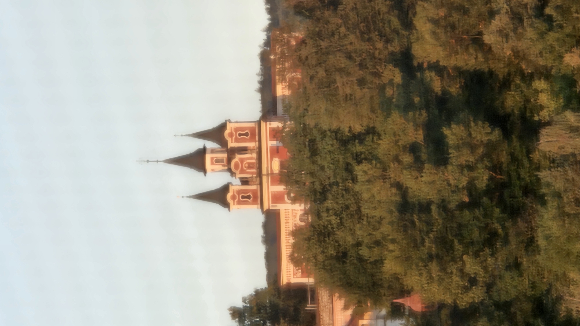 The image shows a castle-like building with three prominent spires, painted in red and white, set against a pale sky. The structure is partially obscured by lush green trees in the foreground. The architectural style suggests a historical or religious building, possibly a church or a palace, with ornate details on its facade and towers. The surrounding environment appears to be natural, with the building perched on a hill overlooking a wooded area. The lighting suggests it might be early morning or late afternoon, with a soft, diffused light illuminating the scene. The overall impression is one of tranquility and grandeur.
The building depicted is the **Saratov Castle** (also known as the Great Saratov), located in the city of Prešov, Slovakia. It is a historic landmark, originally built in the 17th century and later renovated. The castle is situated on a hill overlooking the city and is surrounded by a park and gardens. It is known for its distinctive architecture, which blends Renaissance and Baroque styles, and its picturesque setting amidst nature.