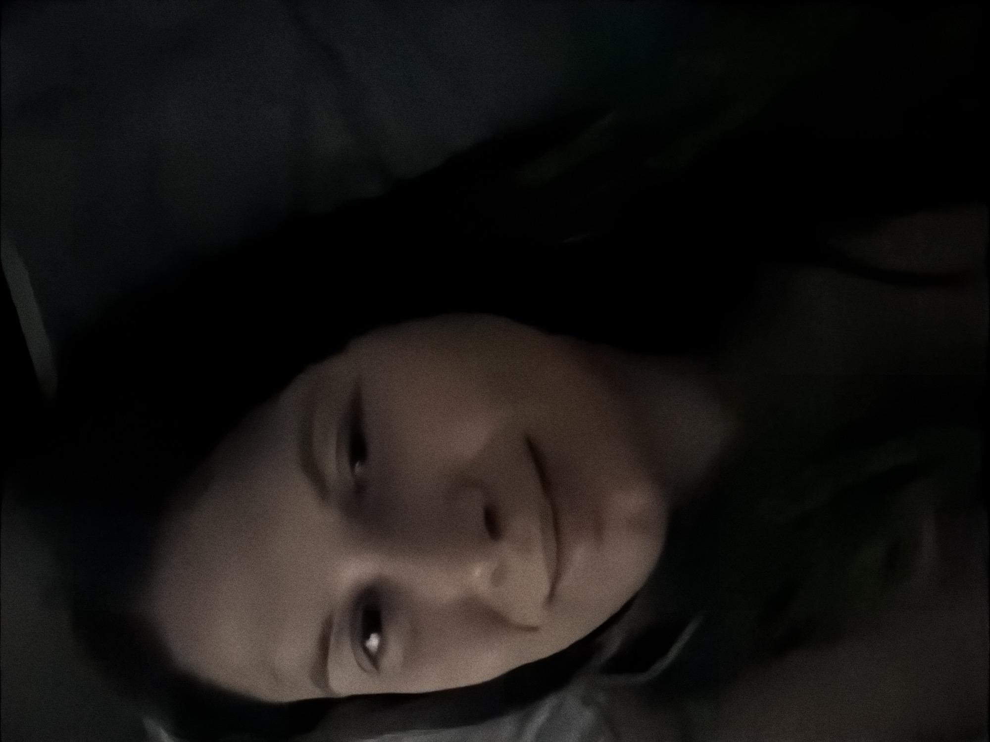 The image is a close-up, dimly lit selfie of a person lying down. The person has long, dark hair and is looking towards the camera with a slight smile. The background is dark and appears to be an indoor setting, possibly a bed or couch. There is no discernible location in the city of Stara Zagora, Bulgaria, visible in the image.