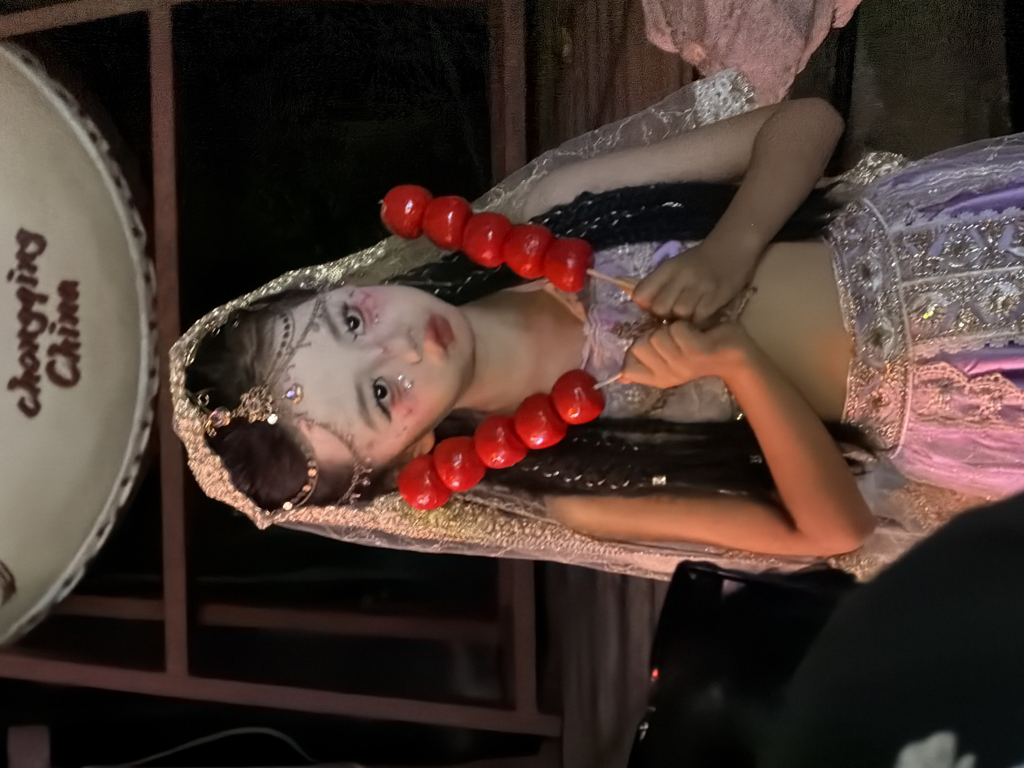 The media file shows a young girl dressed in a purple and silver ornate costume, with a shimmering veil covering her head and shoulders. She is holding a skewer of bright red candied fruit, known as Tanghulu, with a solemn expression. Her face is adorned with elaborate makeup, including subtle glitter and patterned accents. The background is dark and appears to be indoors, possibly a studio or a dimly lit venue, with hints of furniture and equipment. On the left side of the frame, a large, round object with text on it is visible. The text, in a dark cursive font, reads "Chongqing China". The overall scene suggests a performance or a photoshoot, with the girl in costume and the artistic makeup pointing towards a theatrical or celebratory context. The lighting is somewhat dramatic, casting shadows and highlighting the glitter on the girl's costume and face. The orientation of the image is rotated, with the girl's head towards the top and her feet towards the bottom. There are no clear indicators of time of day or weather conditions. The setting appears to be in Yuzhong District, China, as suggested by the visible text.