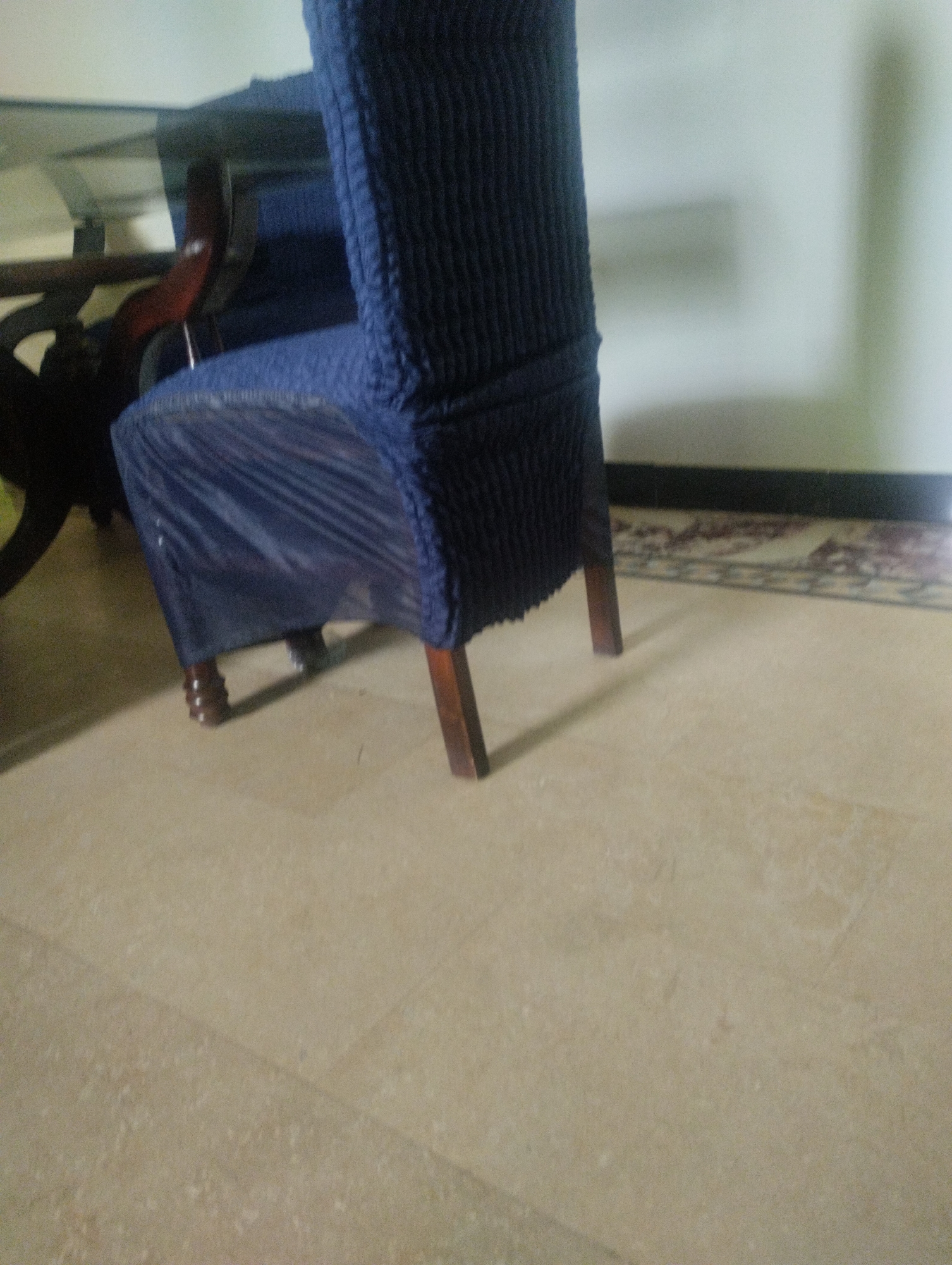 A dark blue chair with a textured surface is partially covered by a translucent blue plastic sheet. The chair has four wooden legs, two of which are visible. The chair is positioned on a light-colored tiled floor. A dark wooden table with a glass top is partially visible to the left of the chair. A carpet with a patterned design is visible in the background, along with a white wall. The scene appears to be an indoor setting, likely a dining room or living area.