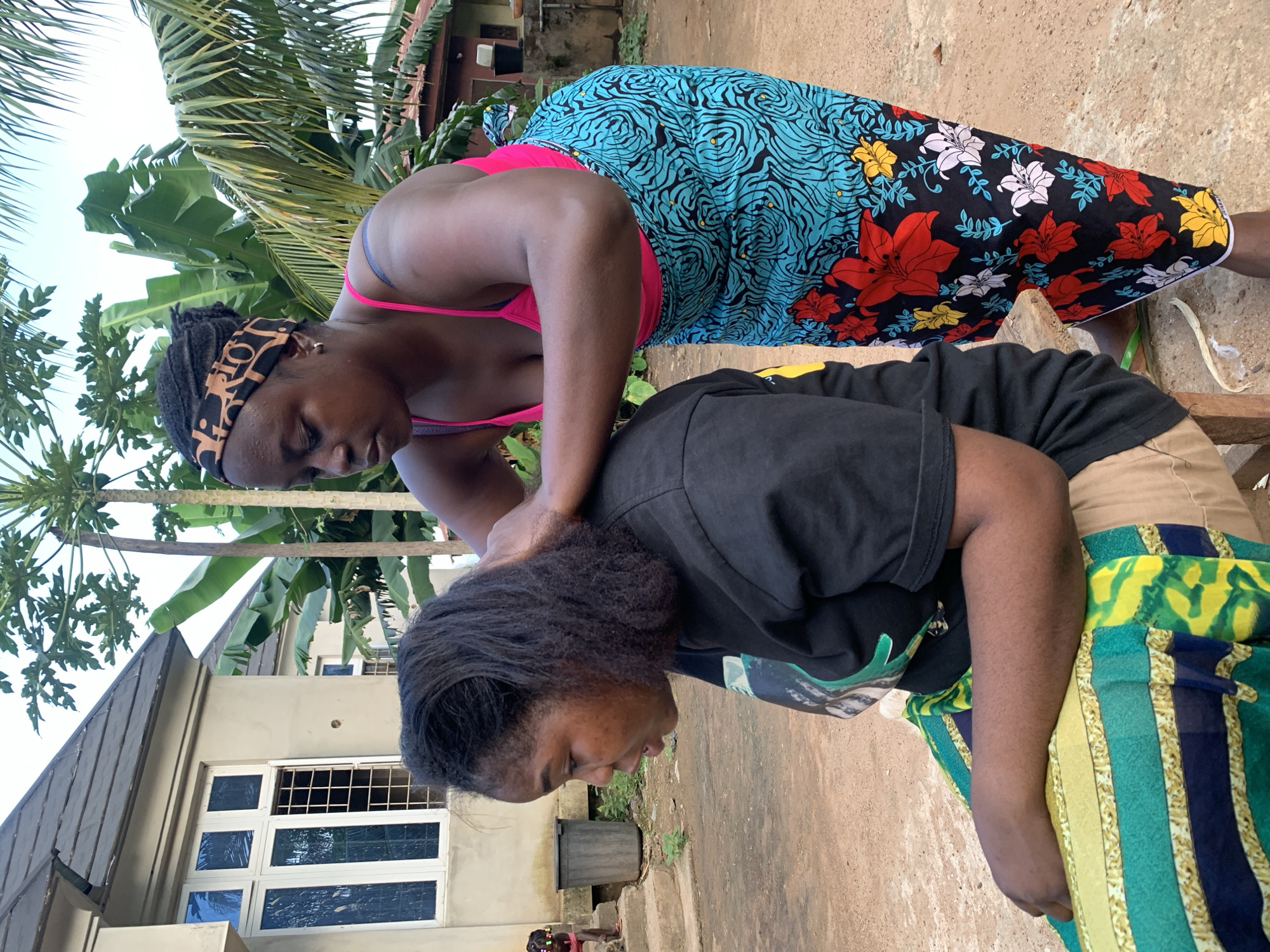 The image shows two young Black women outdoors, appearing to be in a residential area. One woman, wearing a pink bikini top and a colorful patterned skirt, is leaning over and massaging the neck of the second woman. The second woman is wearing a black t-shirt and a green and yellow patterned skirt. She is looking down, and her expression is not clearly visible. The setting includes palm trees and a building with windows. The ground is made of concrete or dirt. The lighting suggests it is daytime, likely mid-morning or afternoon. There is no visible text in the image. The overall mood appears to be relaxed and informal.