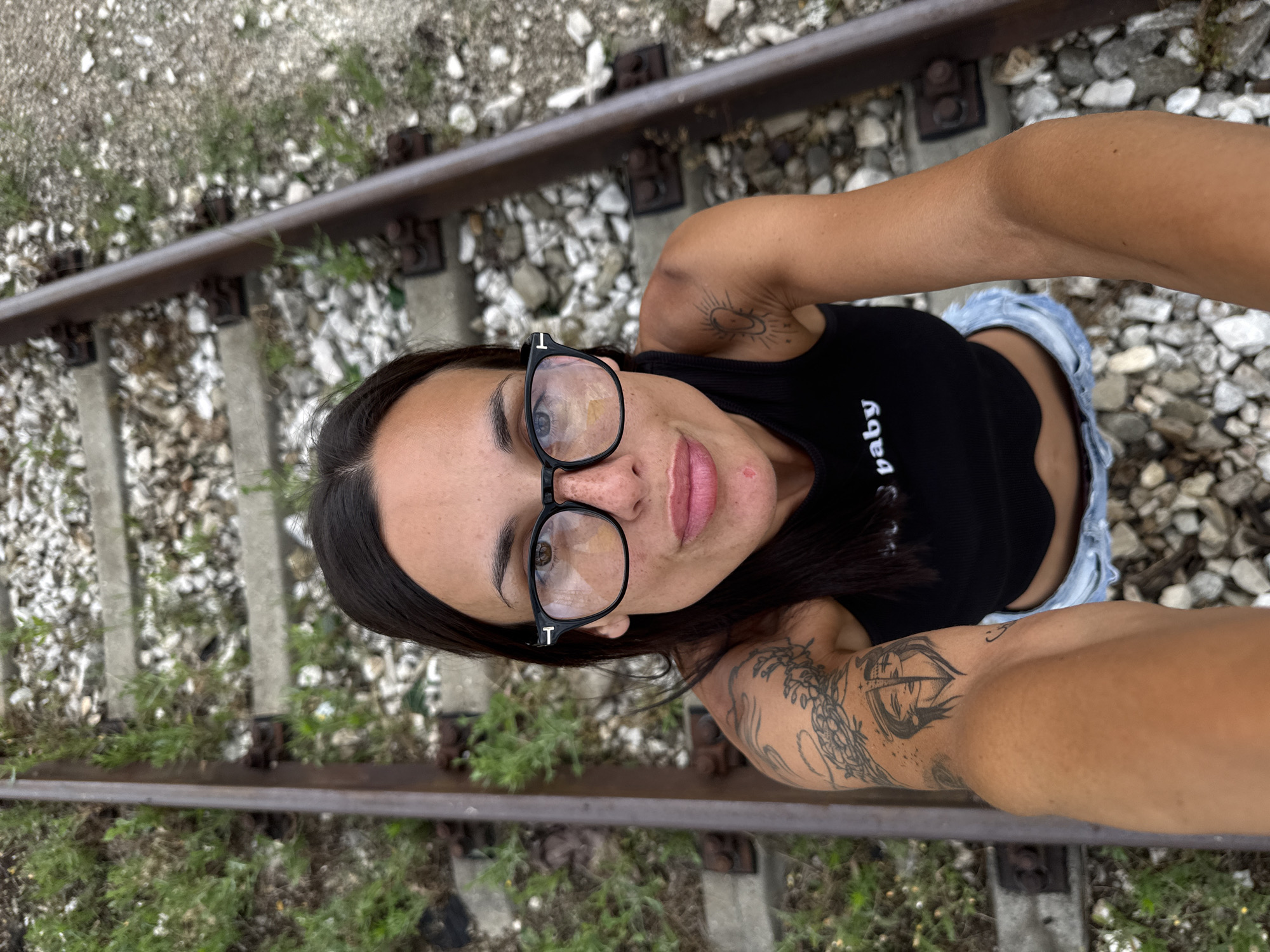 A woman is standing on a railway track, looking at the camera, with her arm extended to take a selfie. She is wearing glasses, a black tank top with the word "baby" on it, and denim shorts. She has tattoos on her left arm and shoulder. The railway tracks are surrounded by gravel and grass. The location is the railway tracks near Pleven, Bulgaria.