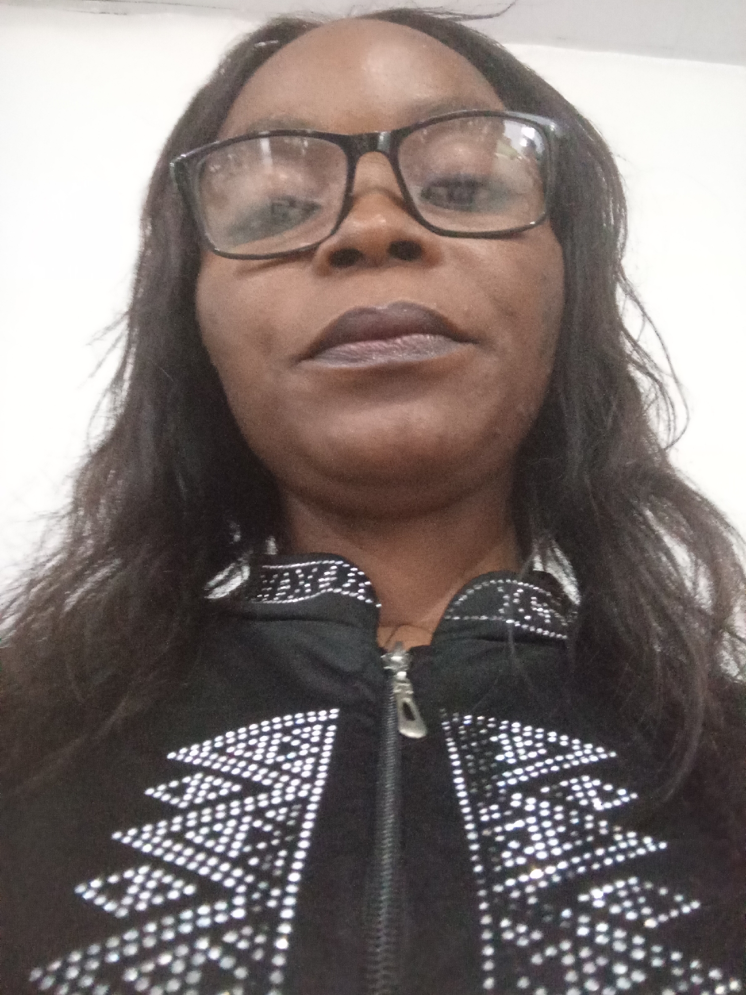The image is a close-up, head-and-shoulders selfie of a Black woman wearing black-rimmed glasses and dark lipstick. She has long, dark hair and is wearing a black jacket with a zipper closure. The jacket is adorned with intricate patterns made of small, reflective studs that form geometric shapes. The background is a plain, off-white wall. The lighting is bright and appears to be from overhead. There is no discernible information within the image that places it in Kason, Nigeria.