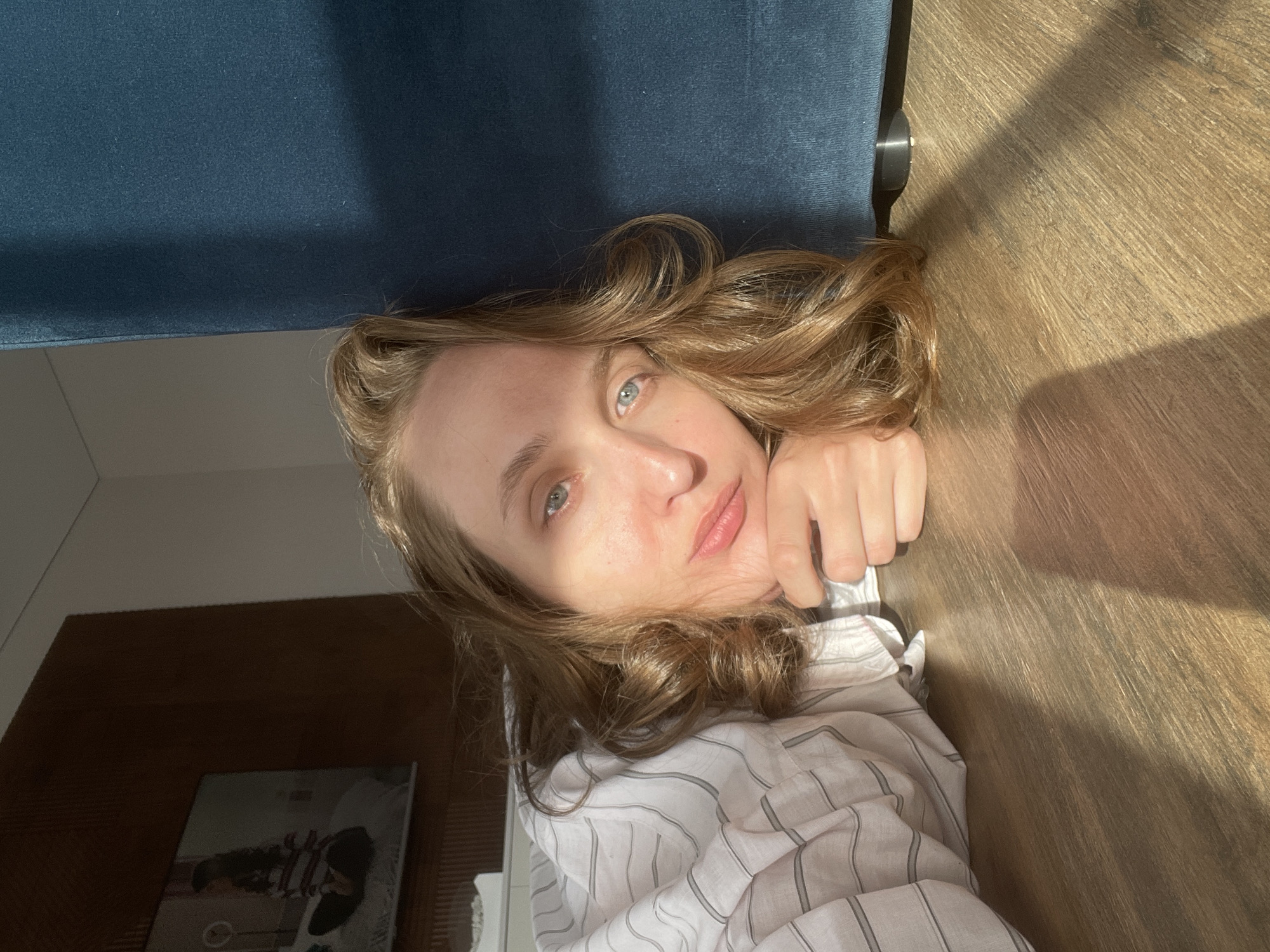 A fair-skinned woman with light brown hair is lying down, partially on a wooden floor and partially on a blue upholstered surface. She is wearing a white and grey striped shirt. Part of a dark wooden cabinet or headboard is visible in the lower left corner of the frame. The setting does not explicitly place the subject in Подшивалово, Russia.
