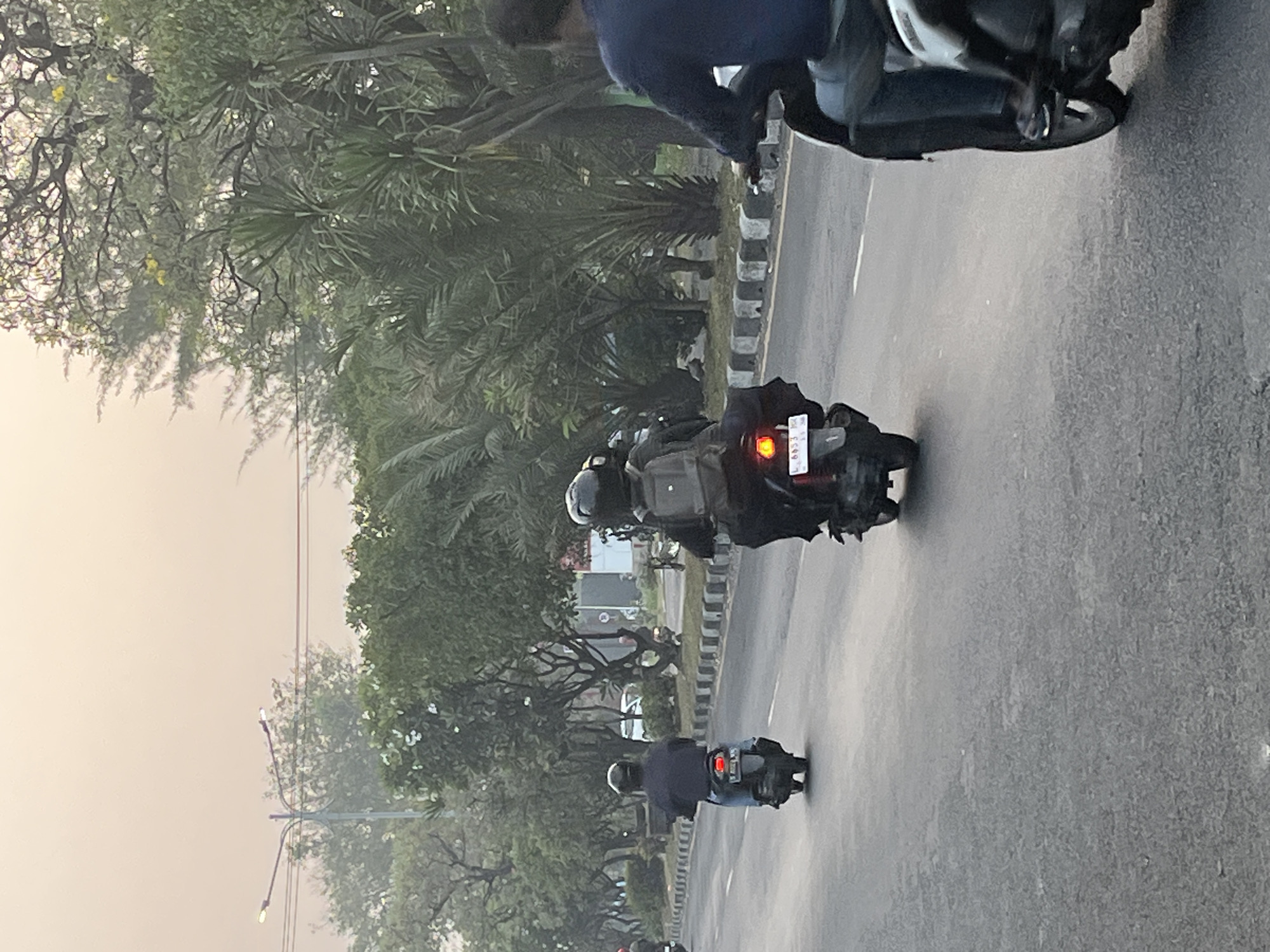 A street scene captures a hazy, possibly overcast, daytime atmosphere with a light sky. The asphalt road has a white lane marker. Several motorcycles are visible, moving from left to right. In the foreground, on the right, a person on a scooter is seen from the side, wearing blue jeans and a dark top. Further down the road, a person on a scooter is viewed from behind, wearing a dark jacket and black pants, with a backpack on. Another scooter is also seen from behind, further to the left, with its rider in dark clothing. In the background, trees with green foliage line the roadside, and a lamppost with two lights is visible on the left. The overall impression is one of a street in a tropical or sub-tropical urban environment. The scene is consistent with a location in Boro, Indonesia, given the prevalent use of scooters and the lush vegetation.