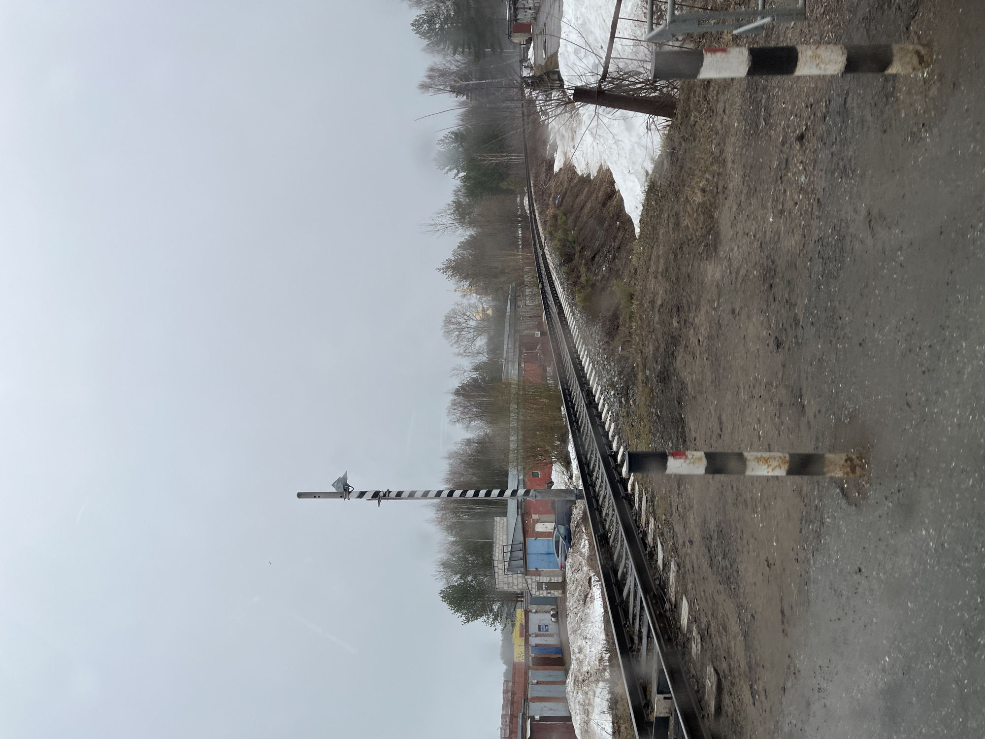 The image depicts a railway track running through an urban or industrial area in Chaikovsky, Russia. The railway is bordered by what appear to be storage units or garages on one side, constructed of brick, concrete, and metal. A striped railway signal is visible. Remnants of snow are visible on the ground. The weather appears overcast or foggy, obscuring the background. No people are visible.