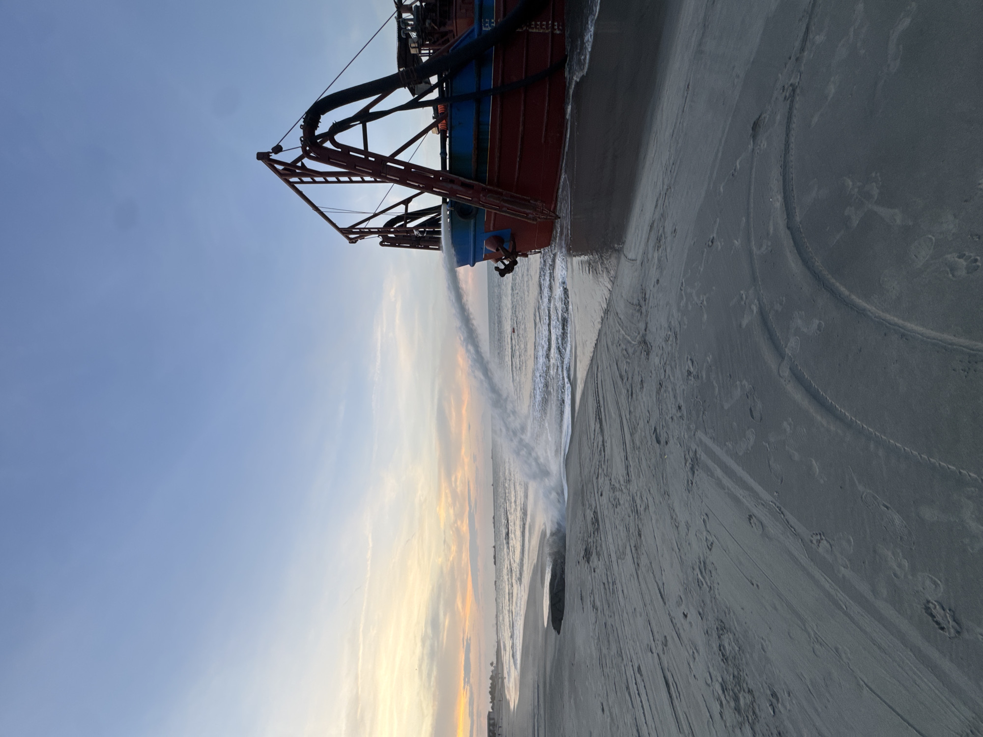 A red and blue ship is beached on a sandy shore. Water is being discharged from the ship towards the shore. The sky in the background is a light blue with wisps of clouds near the horizon. The beach shows tire tracks. Location: Đà Nẵng, Vietnam is not possible to determine.