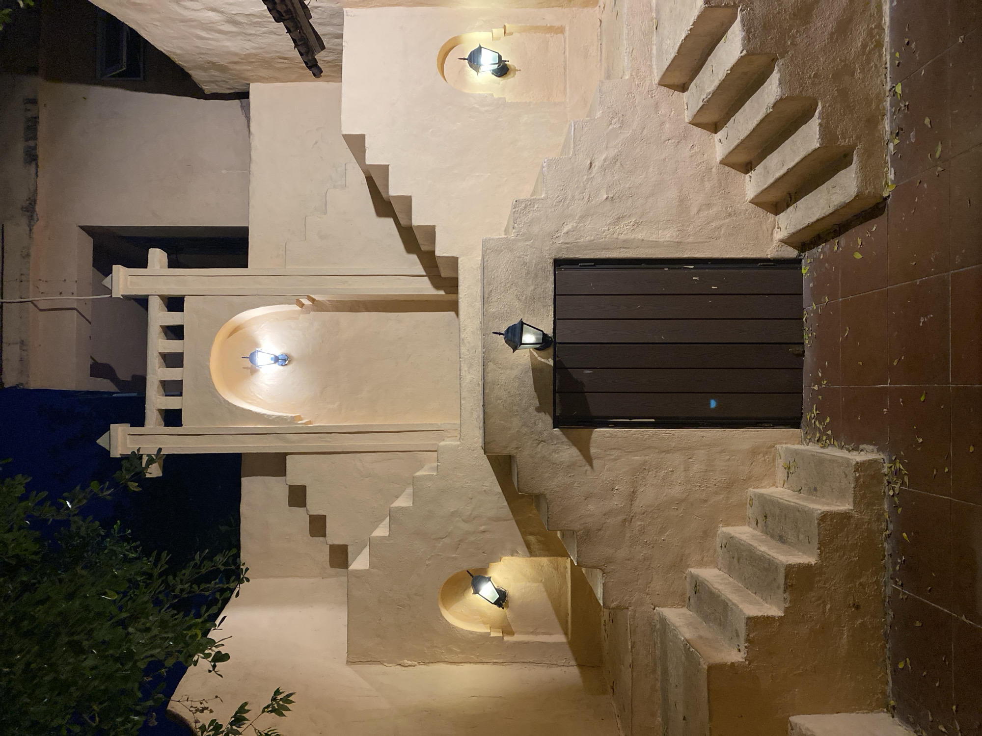 This is an image of an outdoor architectural feature at night, likely a courtyard or pathway. The scene is dominated by textured, pale peach-colored walls and concrete stairs. Several outdoor lamps are mounted on the walls, some within arched niches, casting a warm glow and creating distinct shadows. A dark brown wooden door is centrally located, set within a larger staircase structure. Green leaves are scattered on the ground, which is paved with reddish-brown tiles. A tree with dark green leaves is visible in the upper left corner against a dark blue night sky. The overall aesthetic suggests a Mediterranean or Spanish colonial architectural influence. The image does not contain any people or specific identifiable landmarks that would indicate it is located in the city of Đà Nẵng, Vietnam.