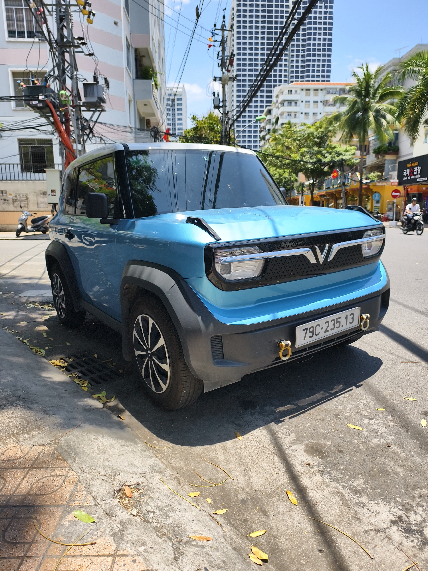 The image depicts a blue VinFast VF 5 electric SUV parked on a city street in Nha Trang Ward, Vietnam. The vehicle is facing the camera, showcasing its front design with the VinFast logo. The license plate number "79C-235.13" is visible. The car is parked on the sidewalk next to a street grate, and fallen leaves are scattered around the base of the car. In the background, there are buildings of varying heights, including a tall, grid-patterned apartment building. Power lines and utility poles are visible overhead. Palm trees and other greenery can be seen along the street. In the distance, a motorbike with two riders is visible near a shop with Korean lettering (NHÀ HÀNG 국제곱창 LÒNG ĐỎ HÀN QUỐC).