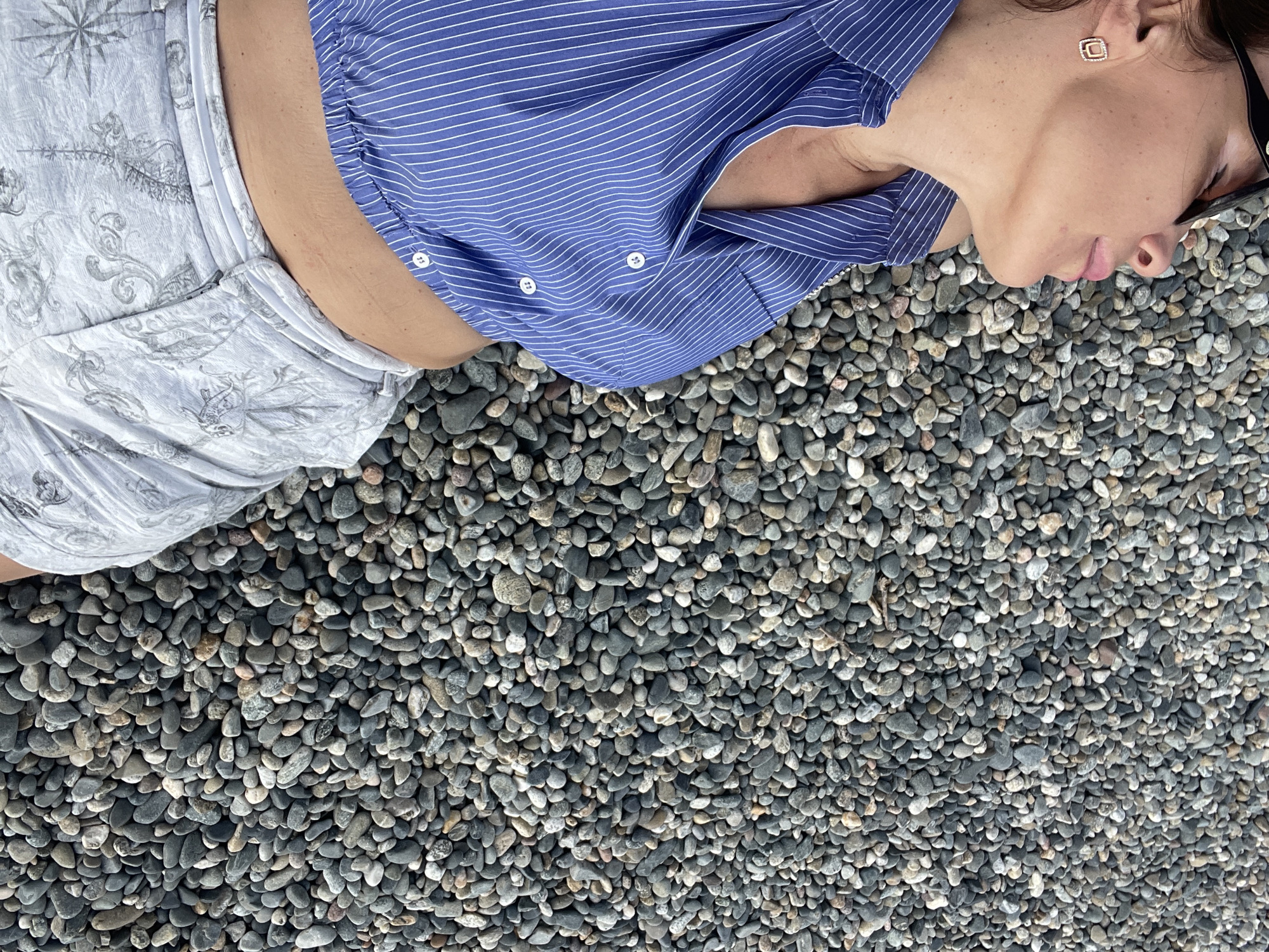 The image shows a person lying on a bed of small, grey pebbles. The person is wearing a blue and white striped shirt and light-colored shorts with a patterned design that includes nautical elements like a compass and anchors. Their head is turned away from the camera, and they appear to be resting or sleeping. The sunlight suggests it is daytime. The setting appears to be outdoors, possibly a beach or a decorative garden area. The pebbles are the dominant visual element of the background, filling the majority of the frame. There is a hint of a person's face, including an ear with a square earring.