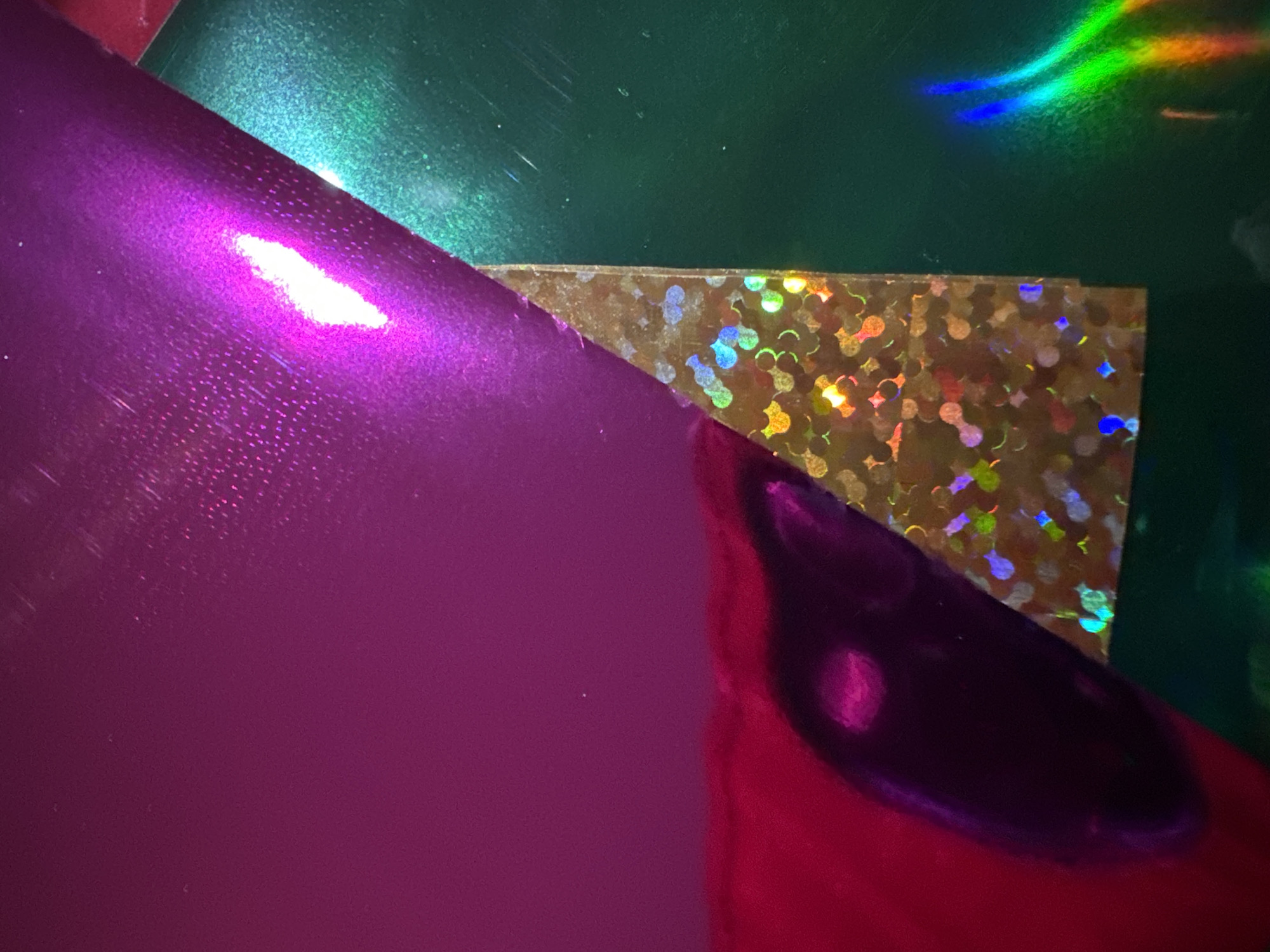 This is a close-up, abstract photograph of overlapping sheets of metallic and holographic paper. The dominant colors are vibrant magenta, deep green, and gold with iridescent holographic patterns. The magenta sheet has a glossy finish with subtle textured effects and a bright white highlight suggesting a light source. It overlaps a dark red material which is partially visible in the lower left corner, reflecting light and appearing somewhat distorted. A section of gold holographic paper, cut into a triangular shape, is positioned between the magenta and green sheets. This holographic paper displays a mosaic of circular and irregular shapes with rainbow-like refractions of light. The deep green sheet, to the right, also has a glossy finish and shows faint scratch marks and some iridescent light reflections. The composition is abstract and does not contain any recognizable elements of the city of Waterford, Ireland.