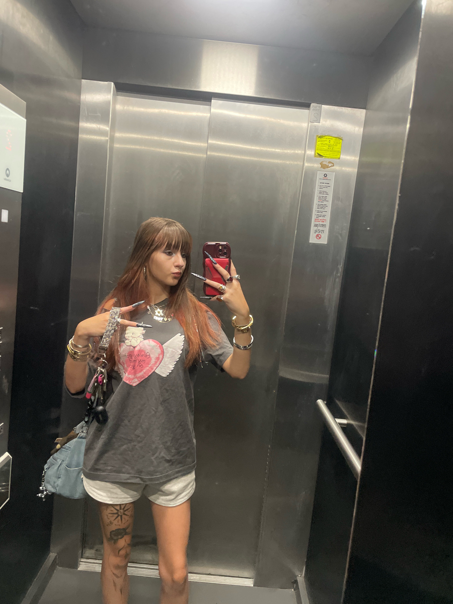A woman is standing in an elevator, taking a selfie with her phone. She is wearing a gray t-shirt with a pink heart and wings graphic, and light gray shorts. She has long brown hair with reddish highlights and bangs, and is wearing several pieces of jewelry, including rings and bracelets. Her fingernails are long and painted dark. She has a light blue denim bag hanging from her left arm, and a keychain with several charms attached to her right hand. Her left leg is visible and has several tattoos. The elevator walls are made of brushed metal, and there are signs on the wall. The floor of the elevator is visible at the bottom of the image. The image is not located in the city of Raanana, Israel.