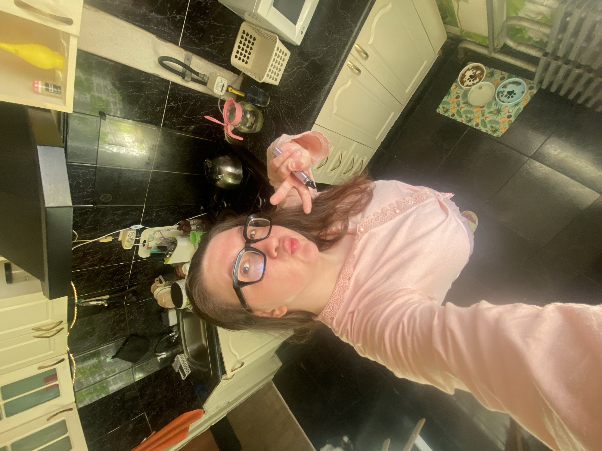 The image displays an adult female in a kitchen setting in Chaikovsky, Russia. The female, positioned slightly right of center, has long dark hair, wears black-framed glasses, and a light pink long-sleeved top with lace trim. She is making a 'duck face' and extends her right hand towards the camera, forming a V-sign with two fingers while holding a dark marker-like object.
The kitchen features a dark, mottled countertop and black tiled walls with a subtle marble pattern, contrasted by light-colored upper and lower cabinets with light brown handles. A stainless steel sink with a faucet is visible on the left, with an orange towel draped nearby. A black wall-mounted utensil holder is positioned above the sink. A silver kettle rests on a stovetop beneath a dark range hood. To the right, a glass pitcher with a pink lid and a small blue cup are on the countertop, alongside a white plastic basket and a white LG microwave oven. A gas meter is mounted on the wall above the countertop. The floor is covered with large dark tiles. On the floor below the microwave, a green mat with a tropical leaf pattern holds three pet bowls: two containing dark material and one with liquid. Artificial strip lighting is present under the upper cabinets, and reflections indicate an external light source.