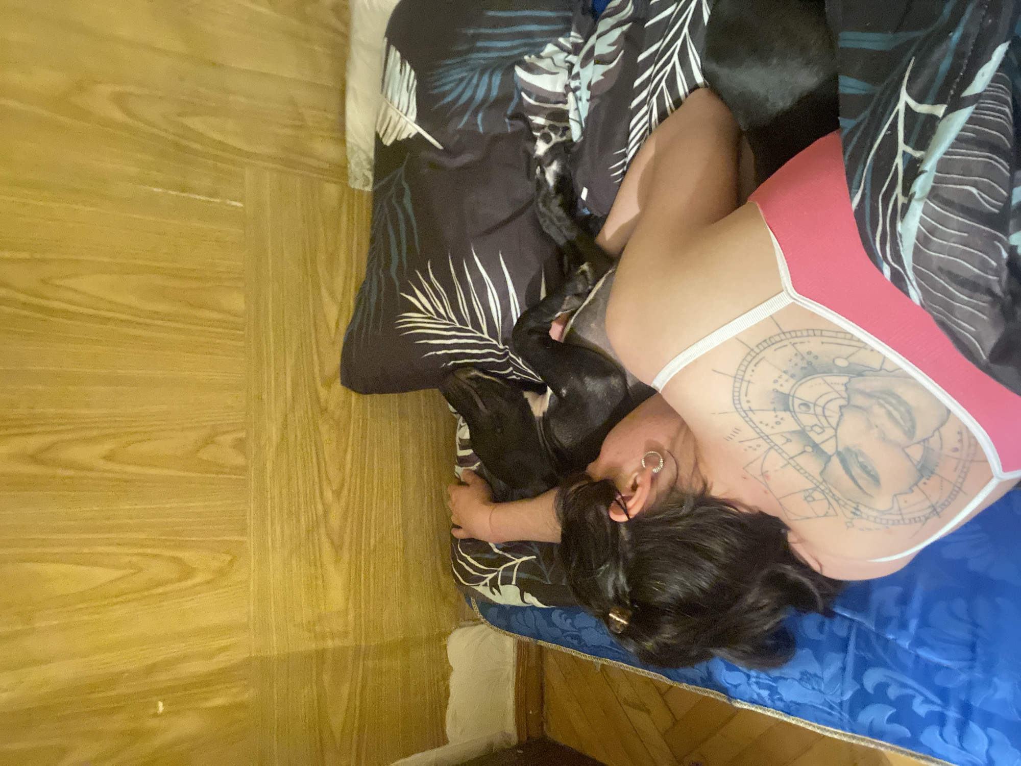 In a bedroom with wood-paneled walls, a person lies on a bed with blue and patterned bedding. The person has dark hair, a tattoo on their back featuring a face surrounded by geometric designs, and is wearing a pink top. A black dog is lying next to the person, partially obscured by the bedding and the person's arm. The scene suggests a domestic, relaxed setting, possibly in a home located in Khimki, Russia.