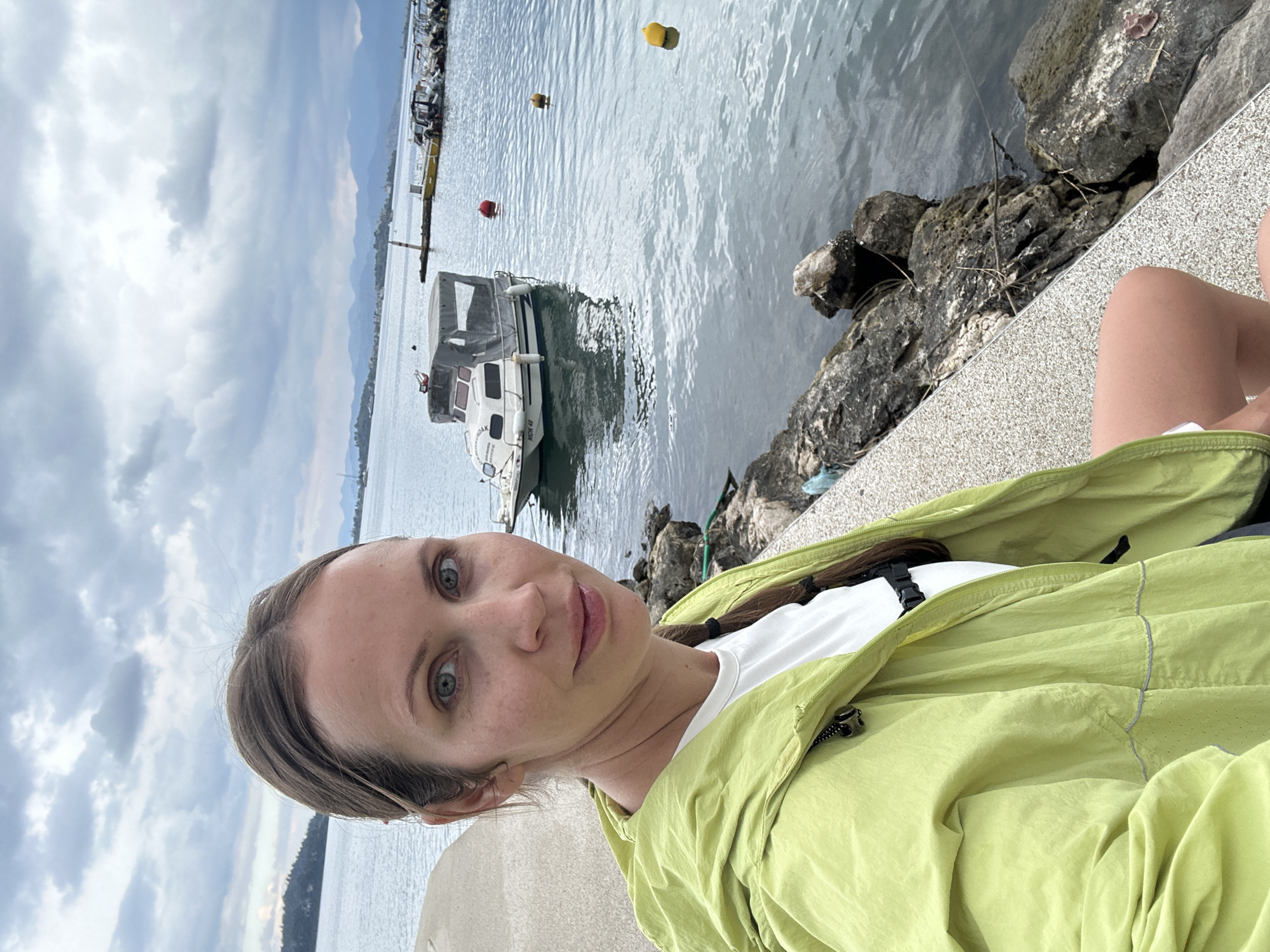 A woman is sitting next to a body of water with boats and buoys, with a cloudy sky above. She is wearing a light green jacket and a white shirt. The location is most likely in Fethiye, Turkey, given the context. The water is near a stone wall, and there are rocks on the edge.
