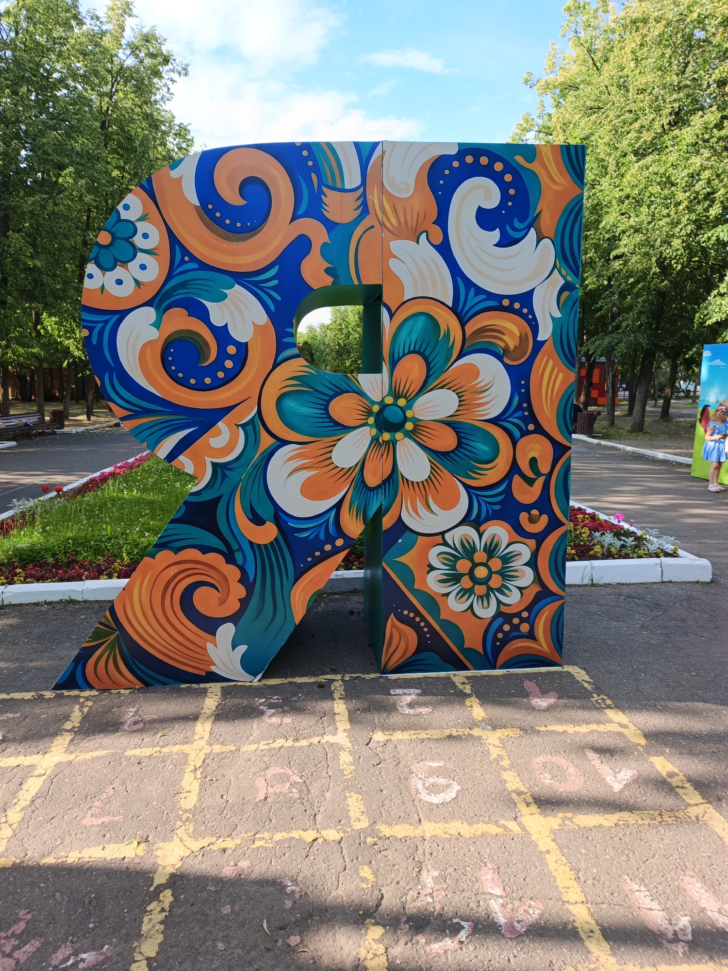 This media file shows a large, colorful letter "R" painted in a folk art style. The letter is decorated with intricate patterns of blue, orange, and white swirls and flowers. It stands outdoors on a paved surface marked with yellow hopscotch squares and faded numbers. In the background, there are trees with green foliage and a blue sky with scattered clouds, suggesting it is daytime and the weather is pleasant. A flowerbed with colorful flowers is visible in front of the letter. Further in the background, a person in a blue dress and a child can be seen near another colorful display, hinting at a public park or an outdoor event. The overall impression is bright and cheerful.