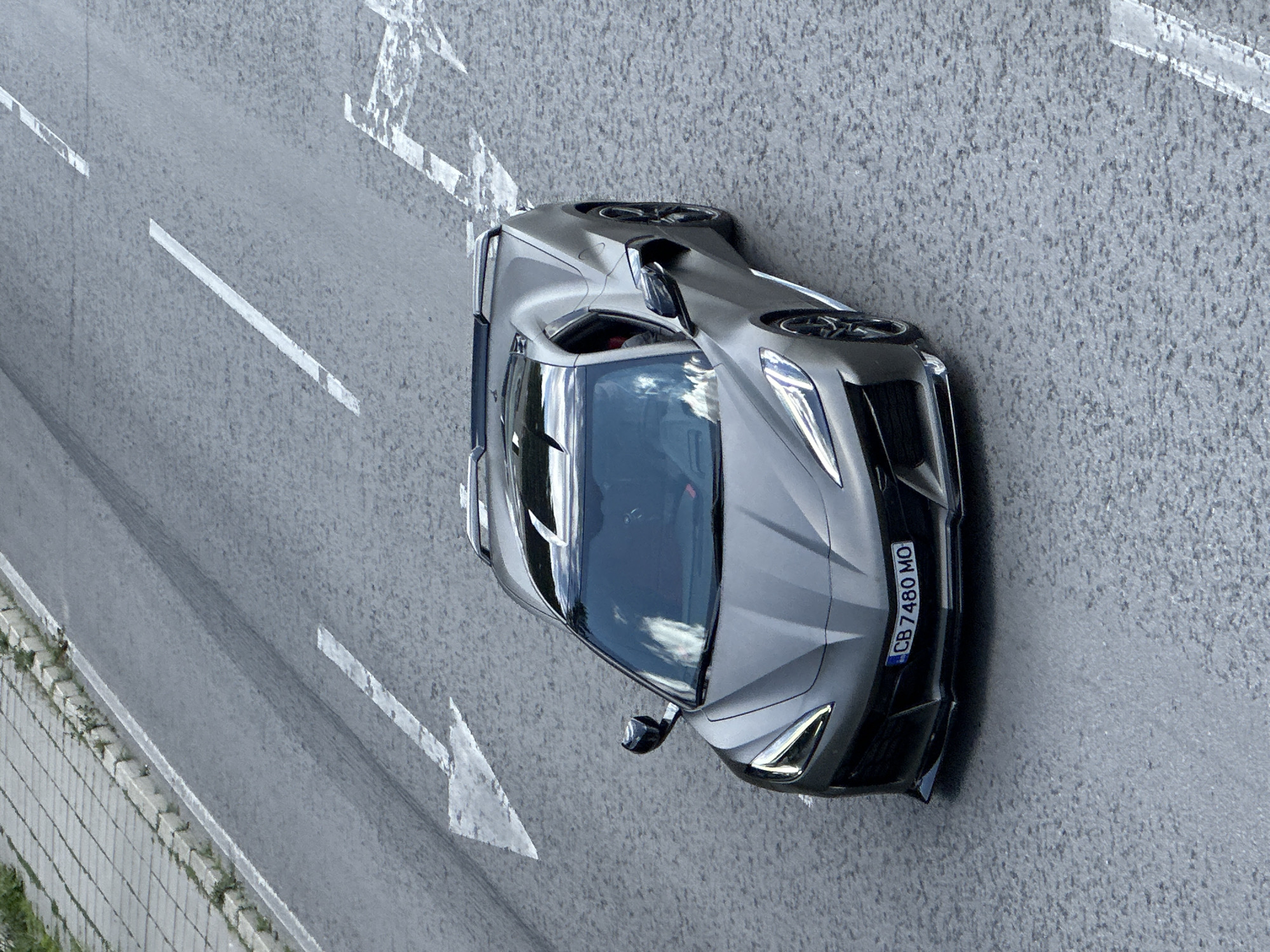 An aerial, eye-level shot captures a dark grey 2020 Chevrolet Corvette C8 convertible driving on a paved road. The car is angled slightly to the right, facing forward, with its front license plate reading "CB 7480 MO". The road features white lane markings, including a left-pointing arrow indicating a turn. To the left of the road is a gray concrete sidewalk with a strip of green grass and a low stone border. The car's roof appears to be retracted. Reflections of the sky and clouds are visible on the car's windshield and body. The image does not show any people or specific landmarks to pinpoint the location in Veliko Tarnovo, Bulgaria, beyond it being a paved urban road.