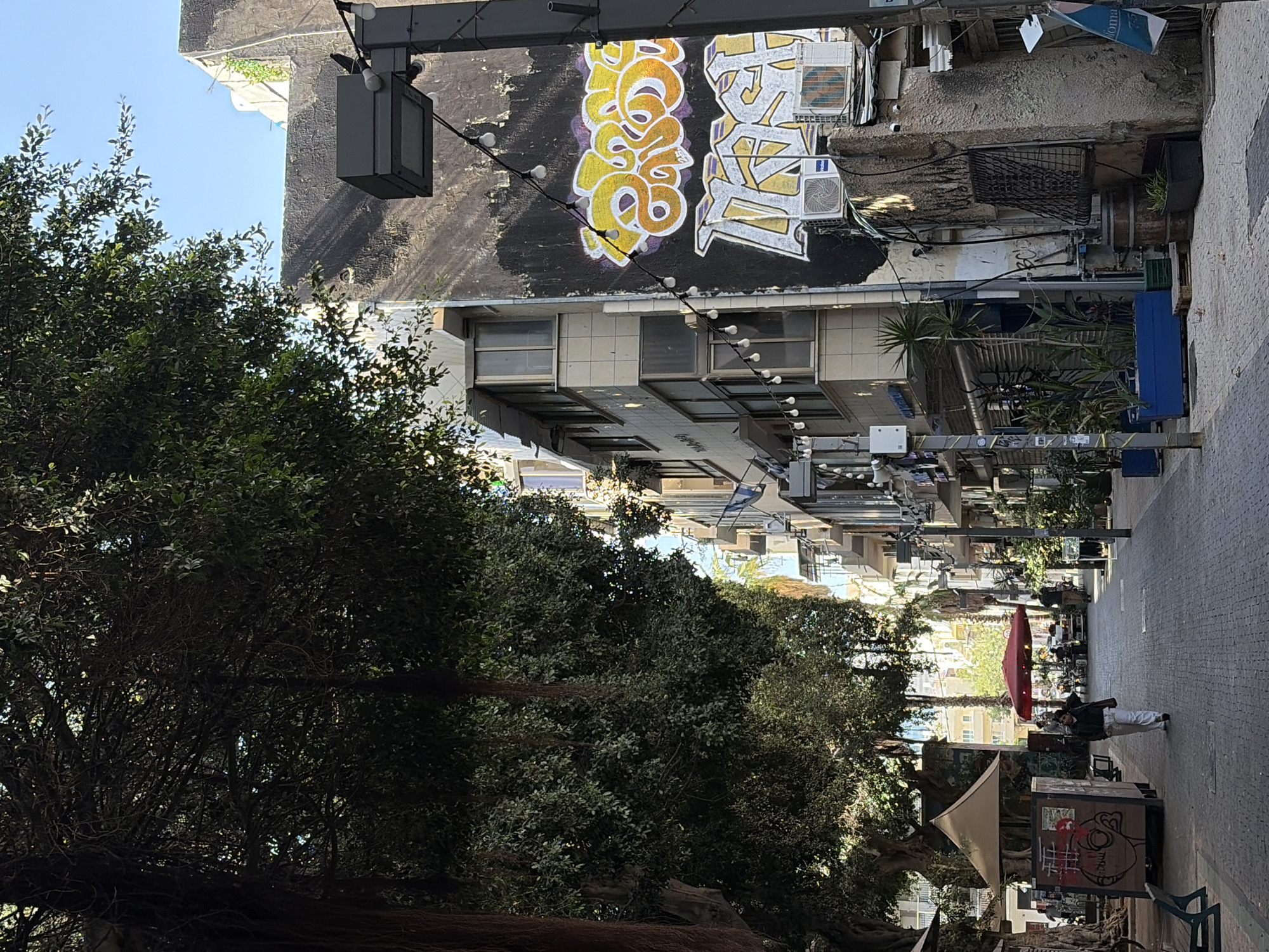 This image captures a street scene in Tel Aviv, Israel, looking lengthwise down the street. The perspective is slightly angled, showcasing the street's buildings and pedestrian area.
Buildings line the street on both sides, with graffiti art noticeable on the higher sections of some structures. Air conditioning units are affixed to the building exteriors. The street is furnished with streetlights, interspersed trees, and pedestrian-friendly furnishings.
A lone individual wearing white trousers and a jacket walks along the street. A red umbrella offers shade to an outdoor seating area for a cafe or restaurant.