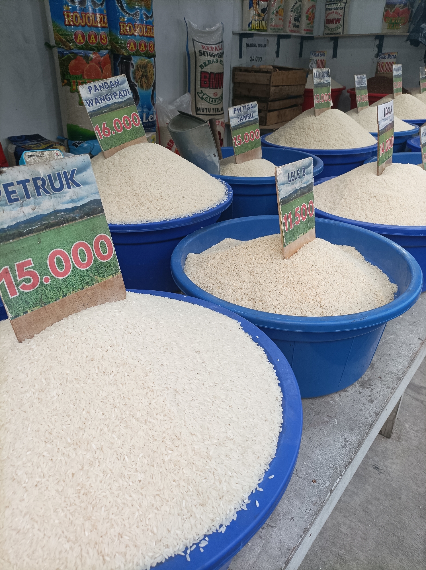 This is a close-up, eye-level shot of a rice stall in a market, likely in West Jakarta, Indonesia, given the context of Indonesian currency and typical market scenes. The foreground is dominated by large blue plastic tubs filled to the brim with white rice. Several price tags are prominently displayed, stuck into the rice. One tag reads "PETRUK" with a price of "15.000". Another, to its right, indicates "PANDAN WANGIPADI" also priced at "16.000". Further back, another tub has a tag labeled "PW TIGA JAMBU" with a price of "15.000". To the right, a tag for "LELE B" shows "11.500", and another for "IDOLA" reads "14.000". Behind these tubs, shelves display various packaged goods, including bags of rice with colorful labels. The background shows a white wall and more shelves with products. The overall environment suggests a bustling, local market setting where rice is sold in bulk.