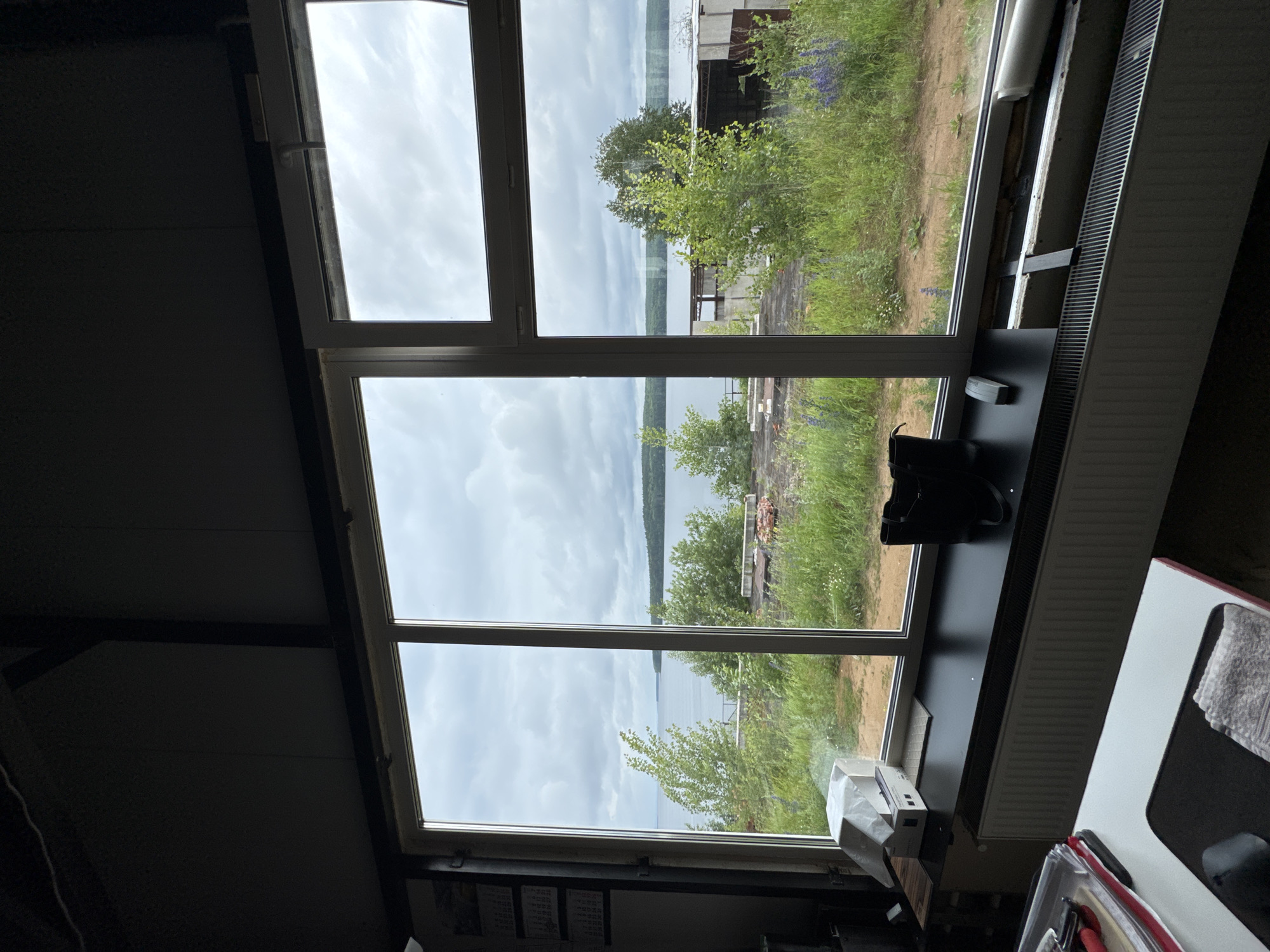 The image depicts a view from inside a building, looking out through a large multi-pane window at a natural landscape. The window frame is white, and the glass is divided into three vertical sections and one horizontal section, creating a grid-like view of the exterior.
Outside the window, a body of water, likely a lake or river, stretches to the horizon. The sky is overcast with gray clouds. Dense green foliage and trees line the shore of the water. In the foreground, closer to the building, there is a grassy area with some wildflowers, including purple lupine. Various structures, possibly unfinished or industrial, are visible amidst the greenery, including concrete blocks and corrugated metal roofing.
Inside the building, on the windowsill, there are a few objects: a white box, a black bag, and a small rectangular object. A radiator runs along the bottom of the window. To the left, a desk with papers and a computer mouse is partially visible. The overall lighting suggests it is daytime but with dull weather conditions. The location is identified as Chaikovsky, Russia.