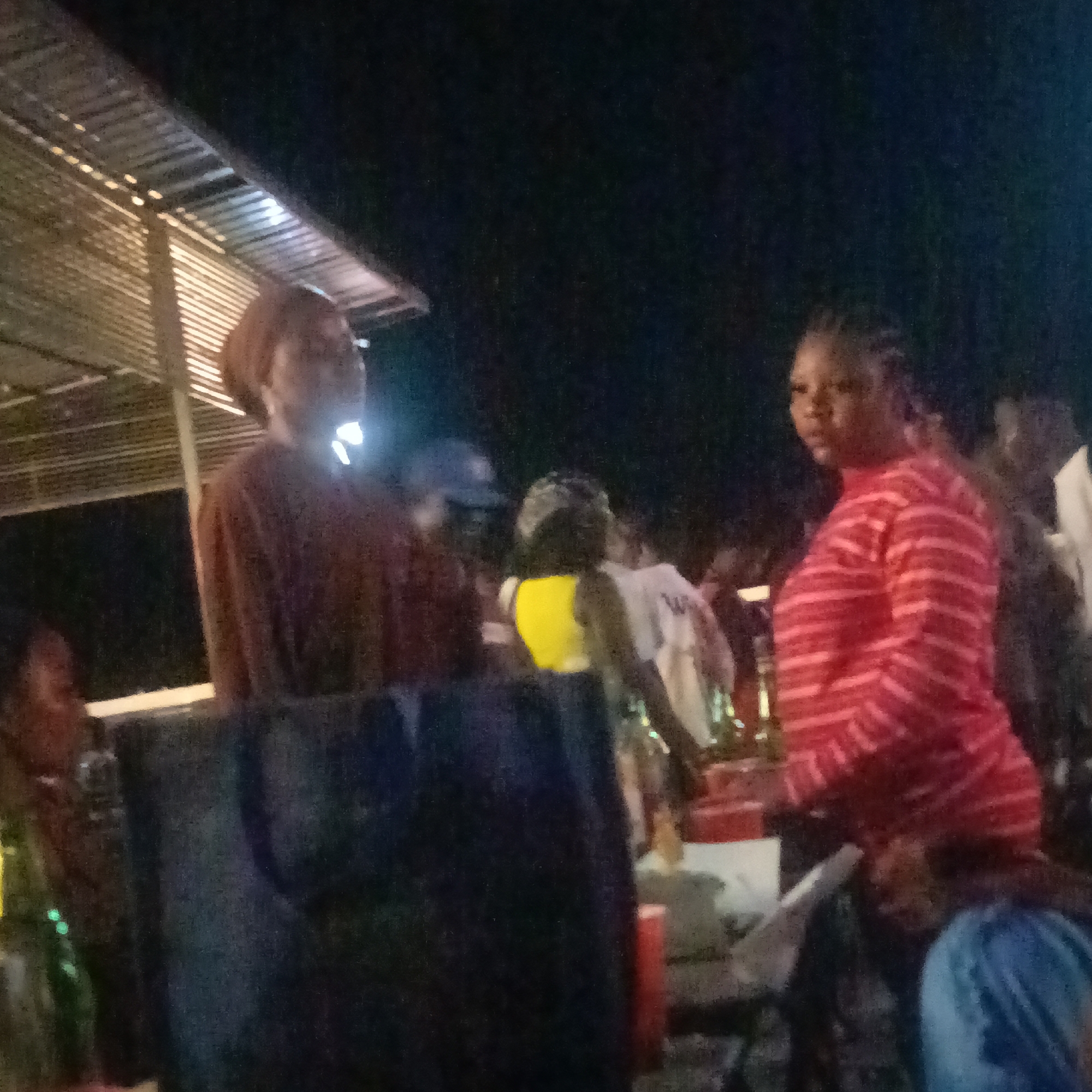 The image, likely taken at night in or near Ube, Nigeria, shows a gathering of people under a corrugated metal roof. Two women are prominent in the foreground: one in a brown top and head wrap, the other in a red and white striped top. Other individuals are visible in the background, some wearing yellow or white tops. There are blurred objects in the foreground, possibly tables or containers, and a green bottle is visible on the left. The lighting is dim, suggesting an outdoor setting at night. The overall impression is of a casual social event or gathering.