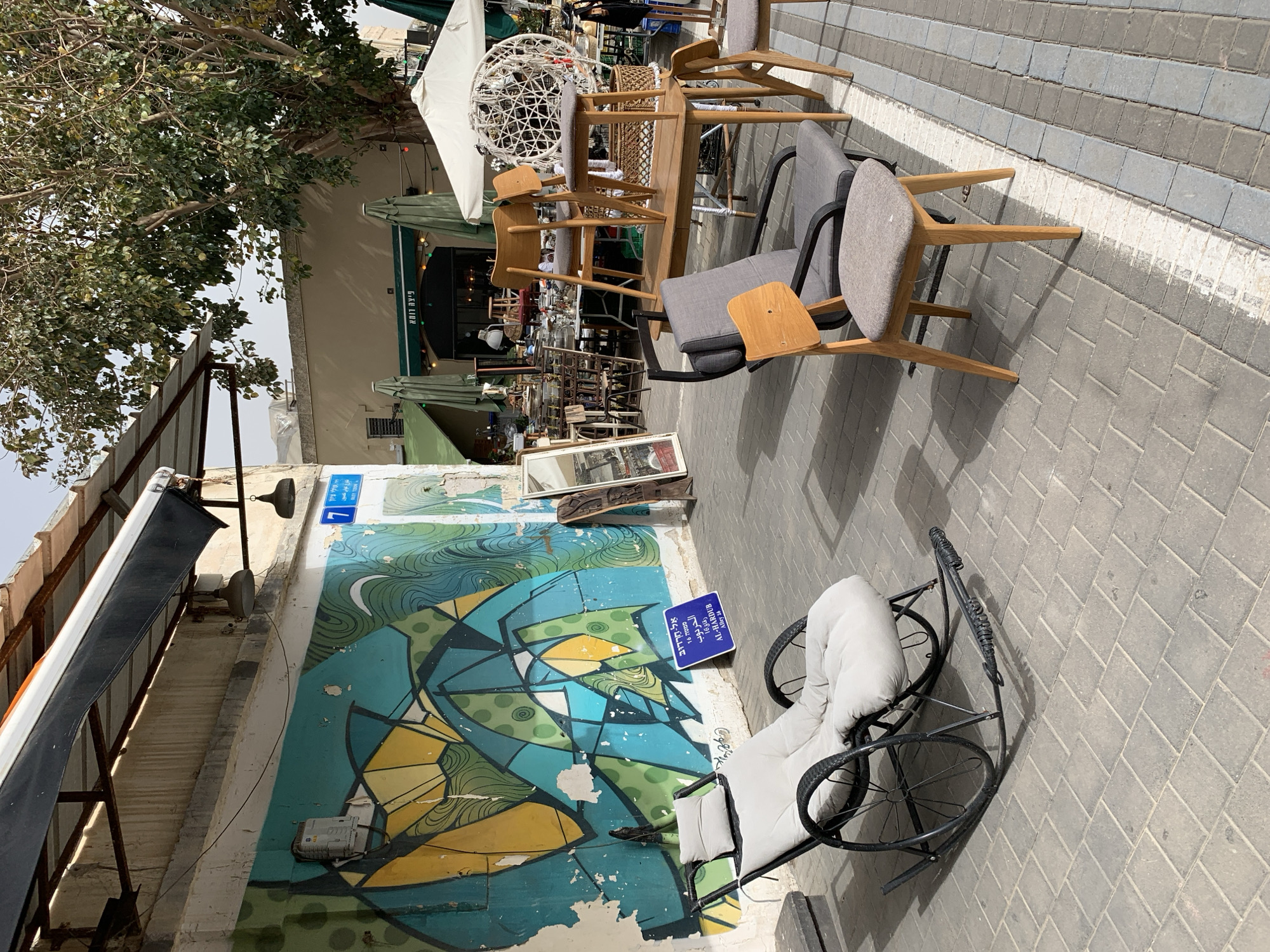 The image depicts an outdoor scene in what appears to be a street in Tel Aviv, Israel, based on the Hebrew signage. A colorful mural covers a section of a building wall, next to which is a rocking chair with white cushions. Next to the wall, there is a blue street sign, partially obscured, that reads "AL-HARDUB Alley 16". The street surface is paved with gray blocks and is cluttered with furniture: chairs, a table, and a hanging chair, all stacked and arranged. Some tables and chairs are in a cafe or restaurant setting with green sun umbrellas. There's a tree providing shade on the left side of the scene. The photograph appears to have been taken during the day. There are no people immediately visible in the shot. The general impression is of a storage area or a street sale of furniture alongside a public area.