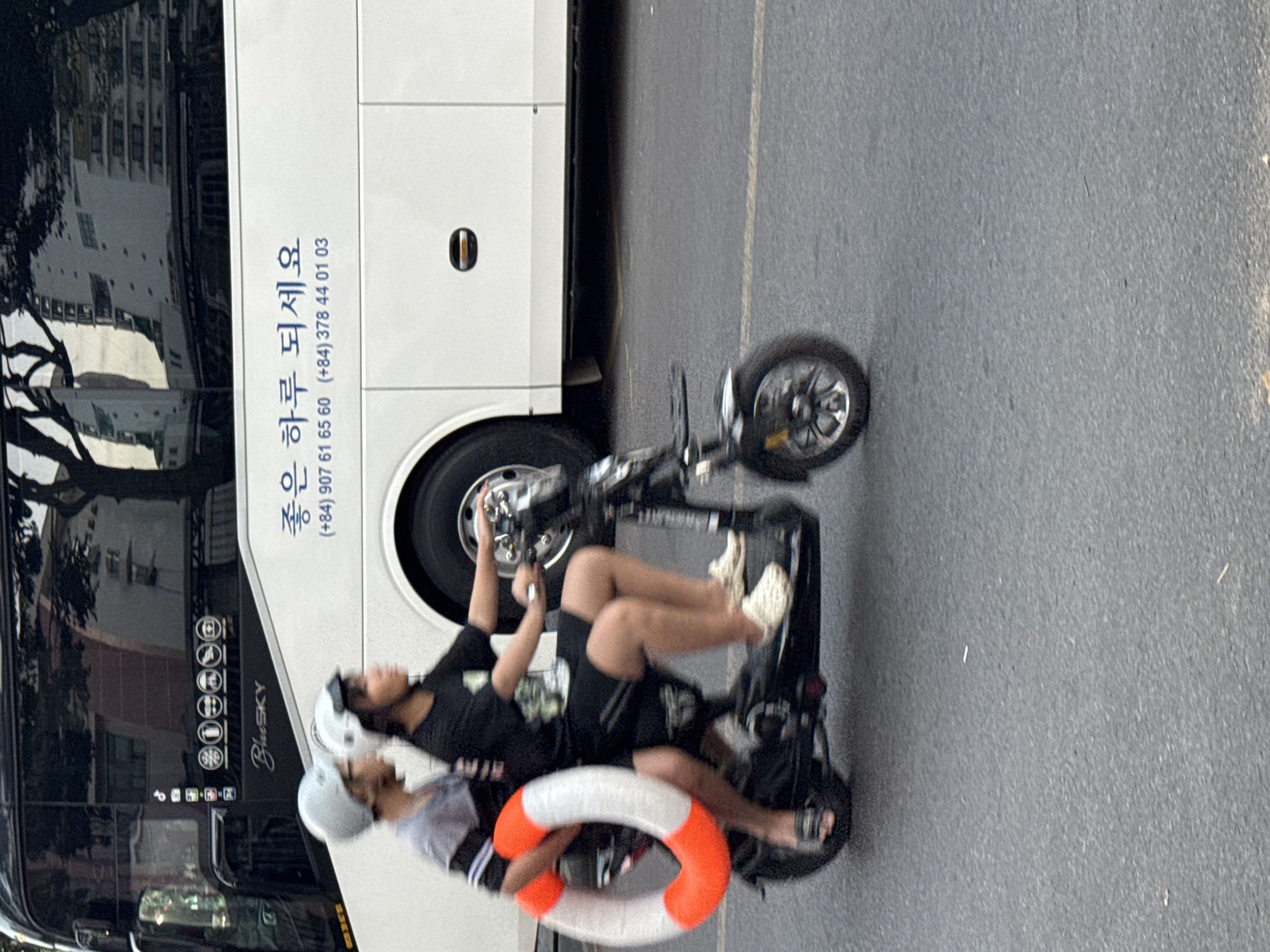 Two young people, one driving a scooter and the other riding on the back, are on a street in Nha Trang, Vietnam. The scooter has an orange and white life preserver attached to the back. The bus behind them has Korean writing on it.