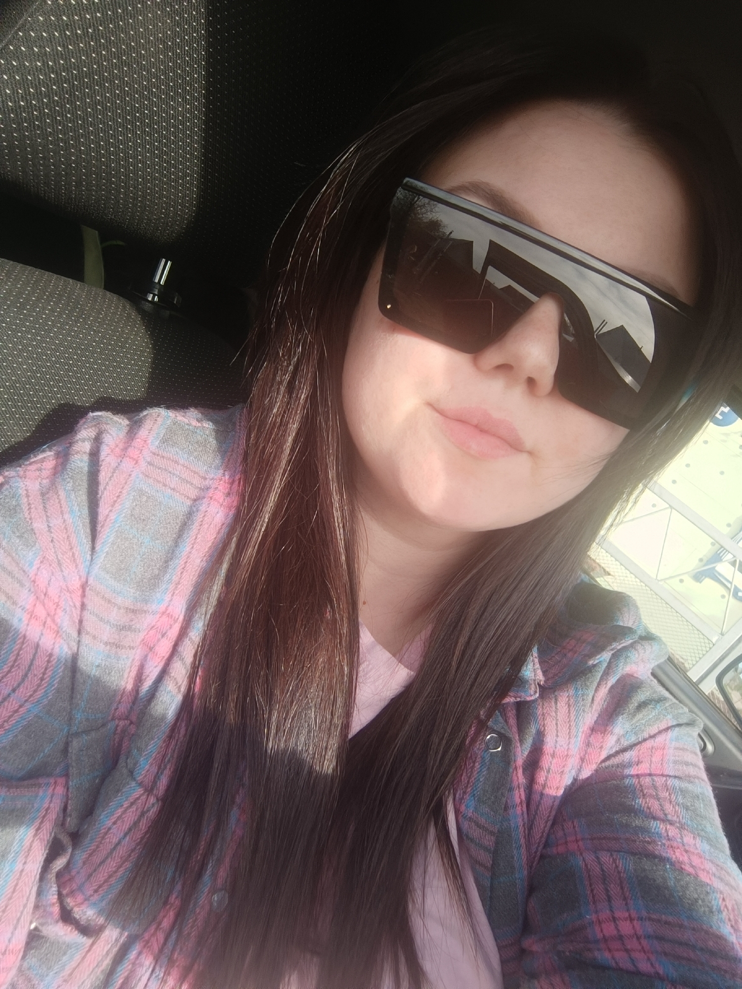 A young woman is taking a selfie indoors, likely in a car. She is wearing large, dark sunglasses, a pink t-shirt, and a pink and grey plaid flannel shirt. Her dark brown hair is long and straight. The car seat behind her is grey fabric with a dotted pattern. The reflection in her sunglasses shows buildings, possibly indicating an urban setting. Without further information, it's impossible to confirm the location is Chaikovsky, Russia.