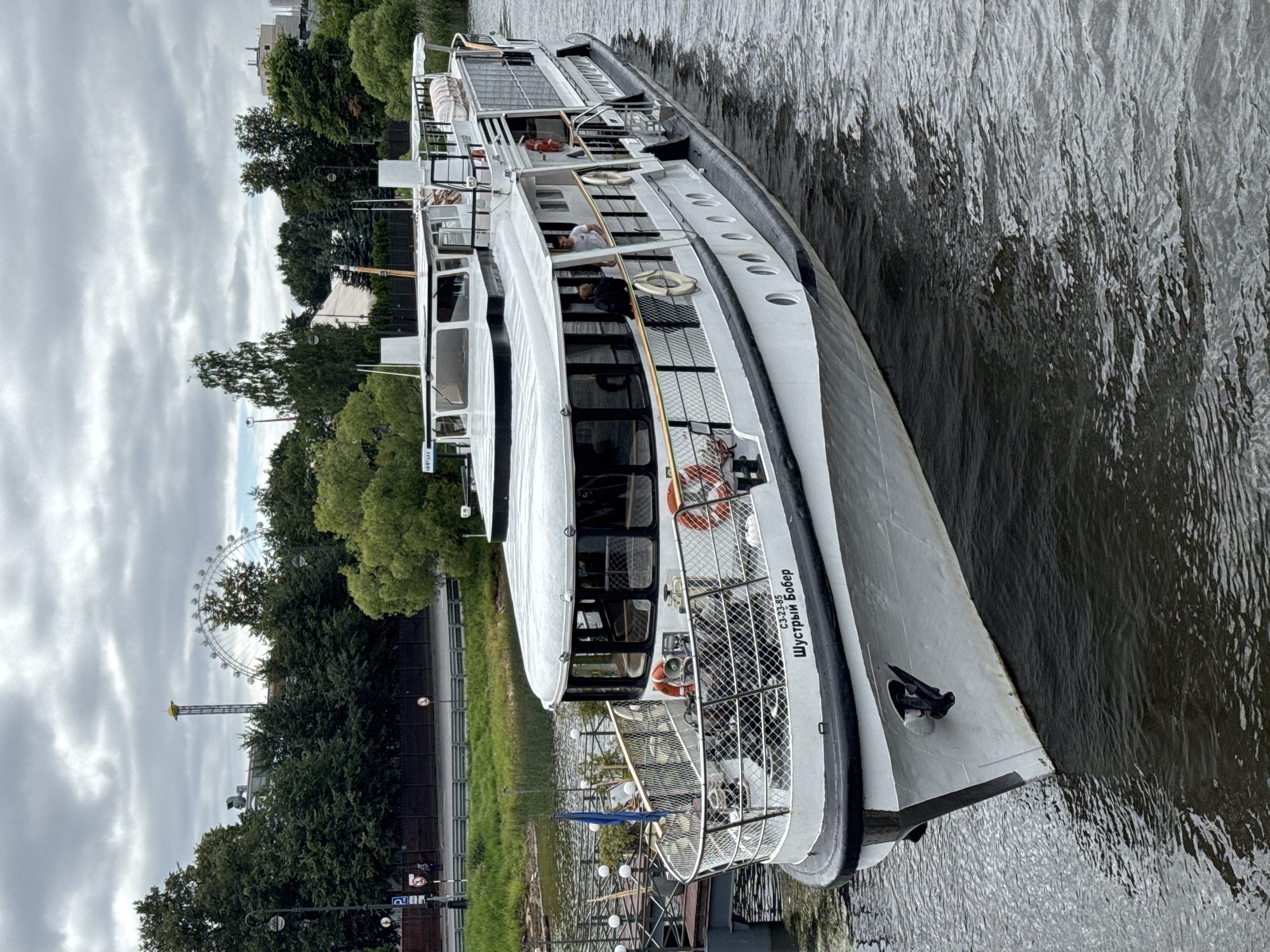 This is a medium-distance, eye-level, medium-shot photograph of a white passenger boat on the Neva River in Saint Petersburg, Russia. The boat, named "ШУСТРЫЙ БОБЕР" (Shustryy Bobr), is white with black trim and features multiple decks, large windows, and a metal railing along the sides. There are a few people visible on the boat, including two men standing on the upper deck and two individuals on the lower deck. The boat is docked at a pier on the left side of the image, with a blue flag flying from a pole. The background shows a lush green embankment with trees, a Ferris wheel in the distance, and a cloudy sky. The water is dark and choppy, with the shadow of the boat cast upon it. The overall impression is one of a leisurely day on the river in a vibrant cityscape.