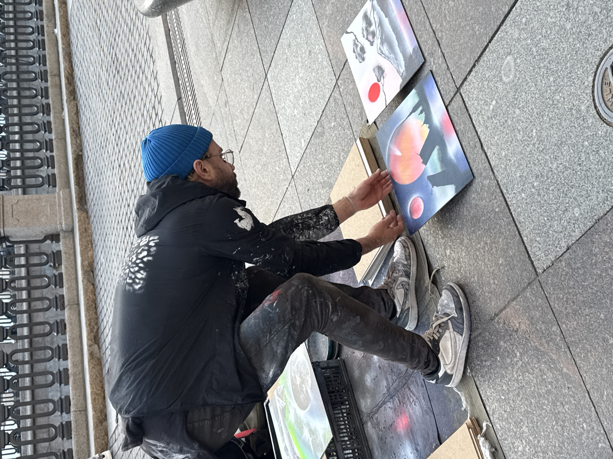 An artist, wearing a blue beanie, glasses, a black jacket splattered with paint, and grey pants also splattered with paint, is crouching on a stone-tiled sidewalk in Saint Petersburg, Russia. The artist is interacting with two paintings laid out on the ground. One painting features a surreal landscape with two moons and a fiery planet. The other painting depicts a traditional Japanese scene with Mount Fuji, dark clouds, and a bright red sun. Behind the artist, a black crate holds what appears to be another artwork, possibly a print with a rainbow or aurora borealis effect. In the background, a decorative metal railing and stone wall suggest a public space, potentially a promenade or embankment. The overall scene captures an artist at work in a public urban setting.
