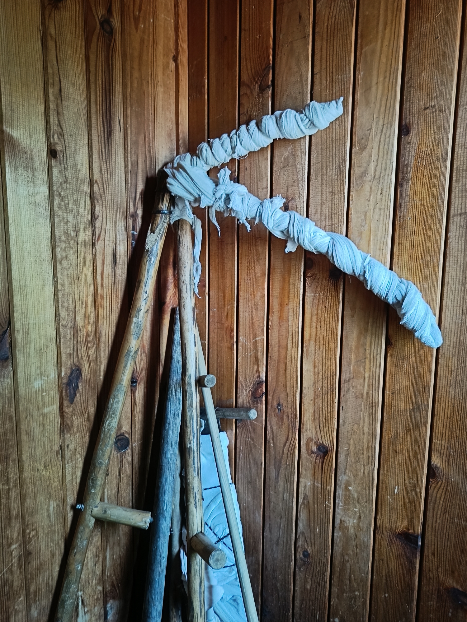A close-up shot captures a collection of wooden tools leaning against a rustic wooden wall. The wall is constructed from vertically oriented planks, exhibiting a natural wood grain with variations in color from light brown to a richer, darker tone. Knots are visible in the wood, adding to its textured appearance.
Dominating the center of the frame is a large, curved object that appears to be a tool or a part of one. It is composed of two wooden poles joined at an angle, with the longer section extending upwards and to the right. This prominent piece is wrapped in a white, somewhat tattered fabric that is twisted around it, creating a textured, rope-like effect. The fabric has frayed edges and is loosely secured, suggesting it might be a protective covering or a decorative element.
To the left and below this main piece, other wooden objects are visible. There's a tripod-like wooden structure with two legs visible, made of rough-hewn wood. A pointed wooden stake, darker in color and with a weathered appearance, leans against the main tool. Another long, thin wooden pole is also present. Some wooden dowels or pegs are attached to the wooden wall itself, serving as hooks. A glimpse of a blue quilted fabric or material is visible behind the wooden objects on the lower right.
The lighting in the scene is somewhat dim, casting subtle shadows on the wall and highlighting the textures of the wood and fabric. The overall impression is one of rustic craftsmanship or perhaps a display of traditional tools in a rural or historical setting.
This scene is located in the city of Semenkovo, Russia, likely within a museum or historical exhibit dedicated to rural life and traditions. The wooden wall and the style of the tools suggest an authentic representation of past agricultural or crafting practices in the region.