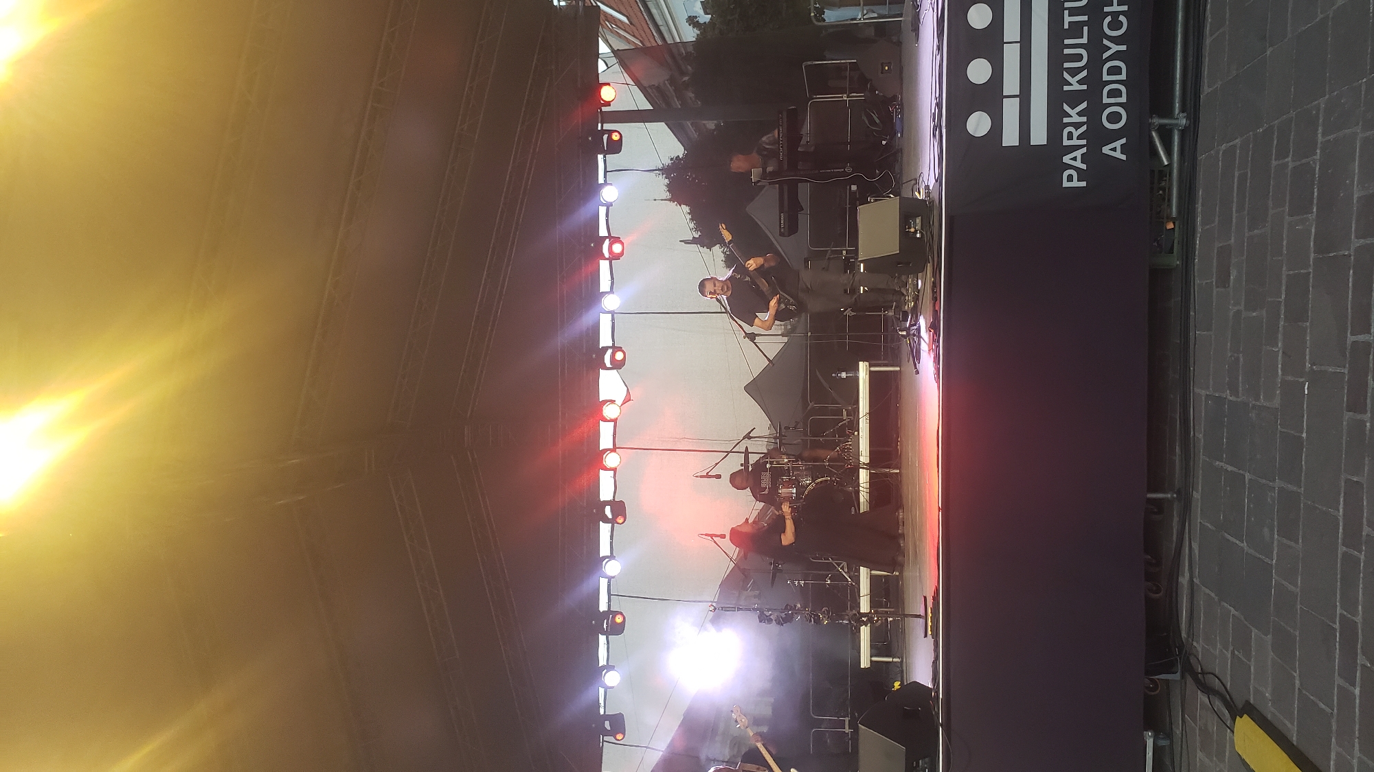 A band is performing on an outdoor stage. The stage is covered by a large tent structure with lighting rigs. The band consists of at least four musicians. One is playing a guitar, another is playing drums, a third is playing a keyboard, and a fourth is playing a bass guitar. A female singer is also on stage, wearing a long black dress. The band is positioned on a raised platform. In front of the stage, there is a large black banner with white text that reads "PARK KULTÚRY A ODDYCHU". The background shows a few buildings and trees. The overall atmosphere appears to be an outdoor concert or event. This scene likely takes place in Prešov, Slovakia, as indicated by the text on the banner.