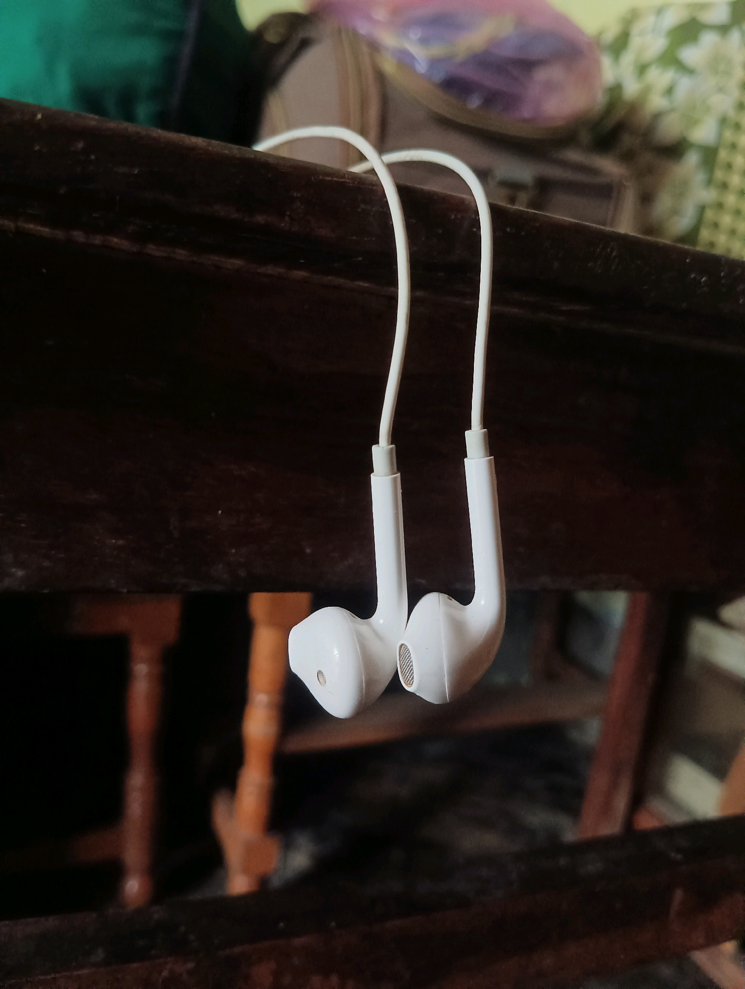 The image displays a pair of white wired earphones hanging from a dark wooden surface, likely a table or shelf. The earphones are positioned in the center of the frame, with their white cables forming a graceful curve as they hang downwards. The earcups are white and have a metallic mesh grille. The background is blurred, showing hints of green and purple fabric, suggesting the earphones are in a domestic setting. The lighting appears natural, casting soft shadows. The specific location in Dakhin Parsa, India, is not visually discernible from the image.