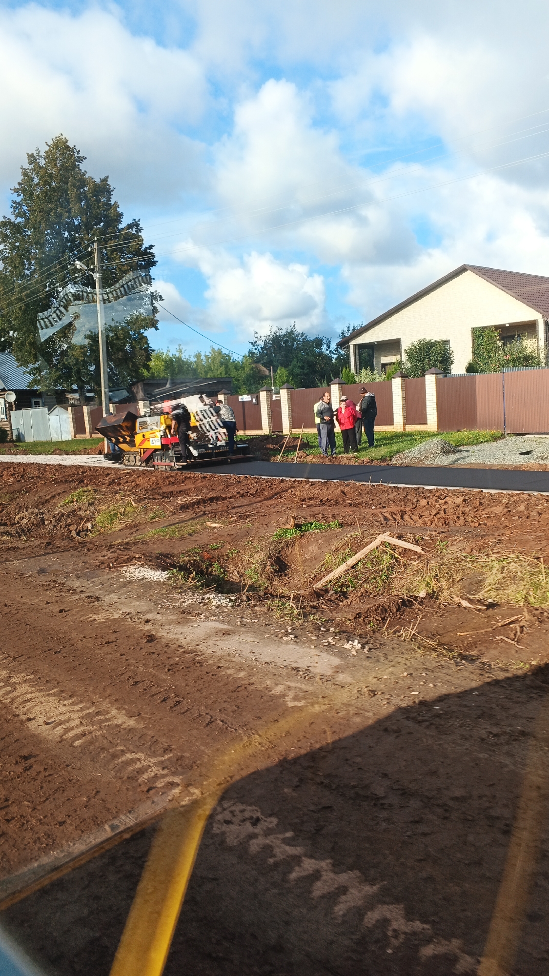In the village of Kiasovo, Russia, an asphalt paver is being operated on a dirt road under a cloudy blue sky. Several individuals are observing the process from a distance. The foreground of the image shows freshly disturbed earth, likely from construction work. In the background, a residential building with a brown roof is visible, alongside a brown fence. The scene appears to be a typical road construction or repair project in a semi-rural setting.