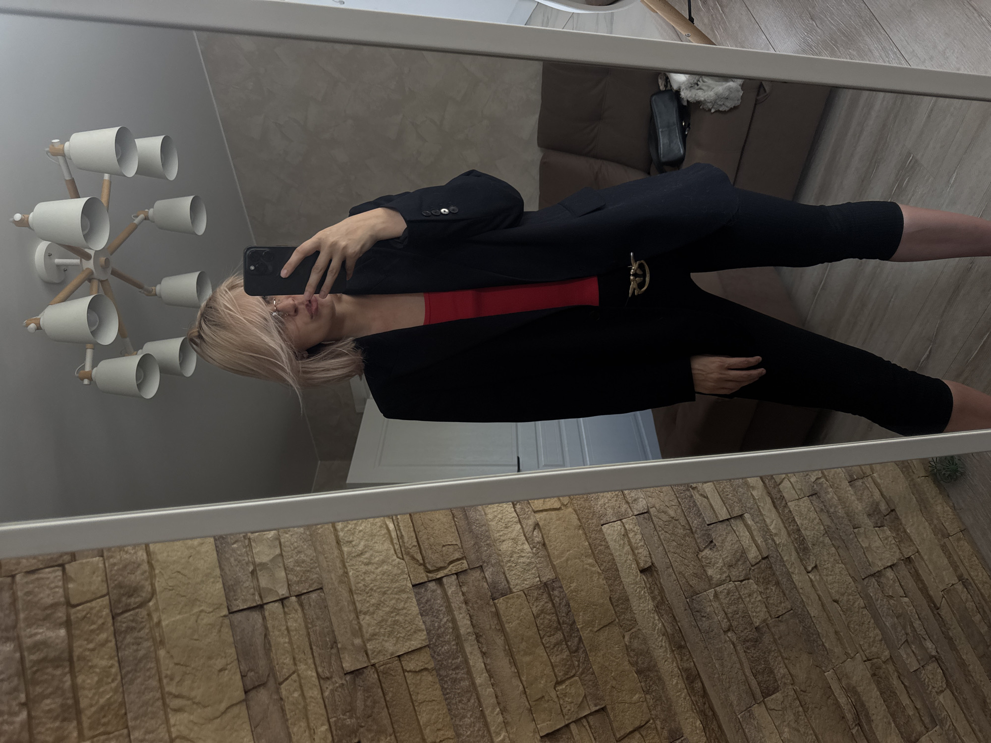 A person with blonde hair is reflected in a full-length mirror, holding a smartphone and taking a picture of themselves. The person is wearing a black blazer with a decorative buckle and a red top, paired with black leggings. The reflection includes a portion of a ceiling light fixture with multiple white shades, a brown sofa with a black bag on it, and white panel doors in the background. The wall adjacent to the mirror is textured to resemble stone or brick. The location may be inferred to be in Saratov, Russia, based on the prompt.