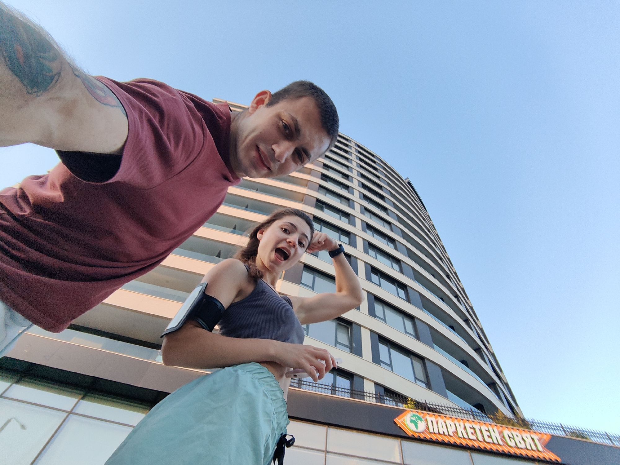 In Varna, Bulgaria, a man and a woman pose for a selfie in front of a modern, curved building. The man, wearing a burgundy t-shirt, angles the camera upwards, capturing his face and shoulder. To his right, the woman, dressed in athletic wear with a phone armband, flexes her bicep, her mouth open in a playful expression. A faint inscription on the building's facade suggests it might be a commercial establishment. The scene is bathed in the warm light of a clear, blue sky.