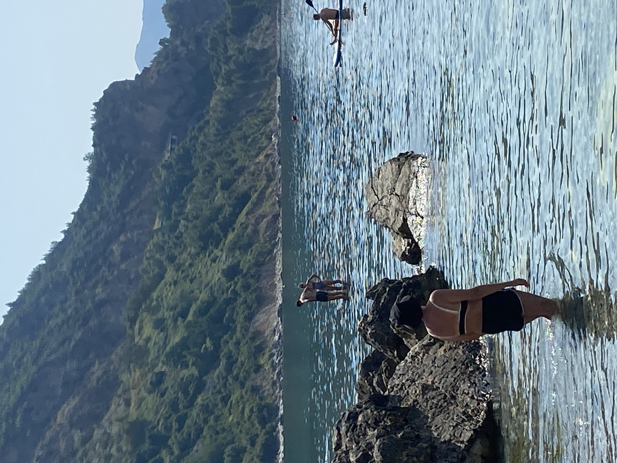 The image depicts a scenic coastal view with a body of water and a lush green hillside in the background. In the foreground, a person wearing a black hat and a black bikini with white straps stands in shallow, clear water, facing away from the camera. The person is partially obscured by large, dark rocks in the immediate foreground.
Further out in the water, a couple, a man and a woman, stand facing each other. The man is wearing dark shorts, and the woman is wearing a bikini. They appear to be interacting, possibly conversing or embracing.
To the right of the couple, a person is seated on a paddleboard, holding a paddle, and is positioned further out in the water. In the middle distance, a small, red buoy is visible in the water.
The environment is characterized by a calm, blue sea with ripples reflecting the sunlight. The background features a steep, verdant hillside covered in dense vegetation, with some rocky outcrops and a very small building perched on its slope, barely visible. The sky above is clear and light blue, suggesting a sunny day. The location is identified as Prijevor, Montenegro.